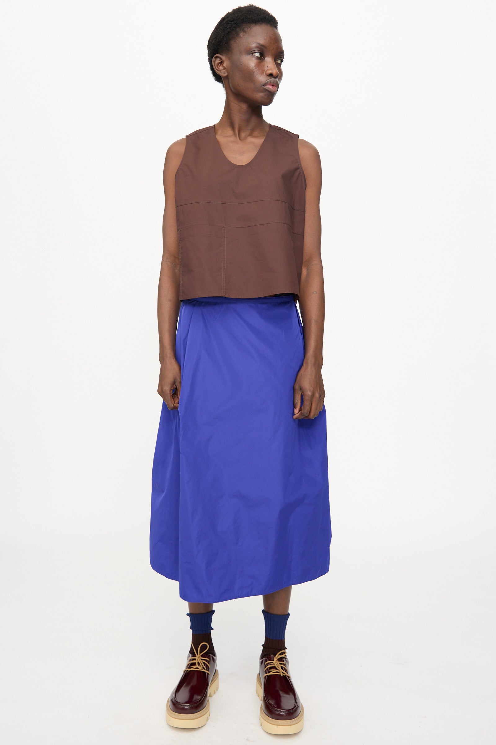 A person stands against a plain background, wearing a sleeveless brown top, blue socks, maroon lace-up shoes, and the Sofie D'Hoore Taffeta Suvi Skirt in Klein.