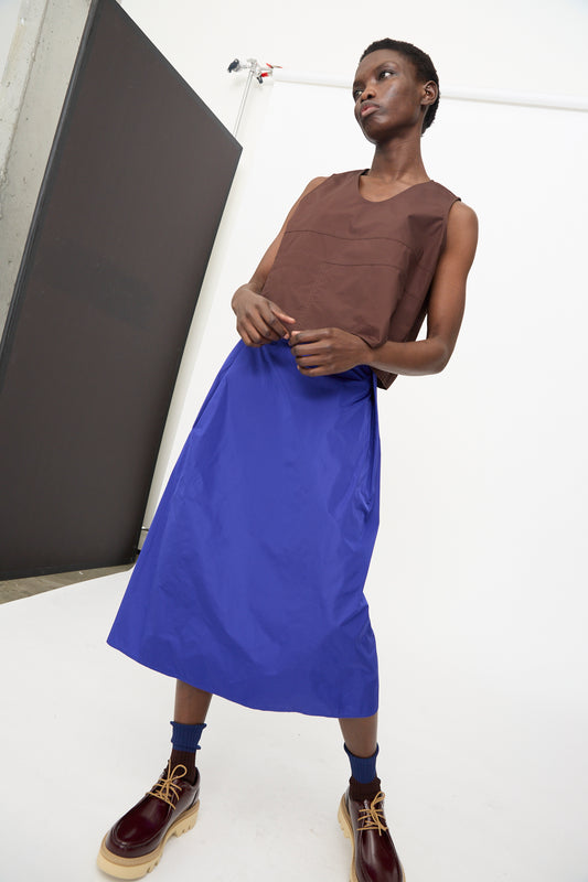 A person stands indoors in a sleeveless brown top, navy socks, and burgundy lace-up shoes with thick soles, wearing the Sofie D'Hoore Taffeta Suvi Skirt in Klein. The background is minimal and white.