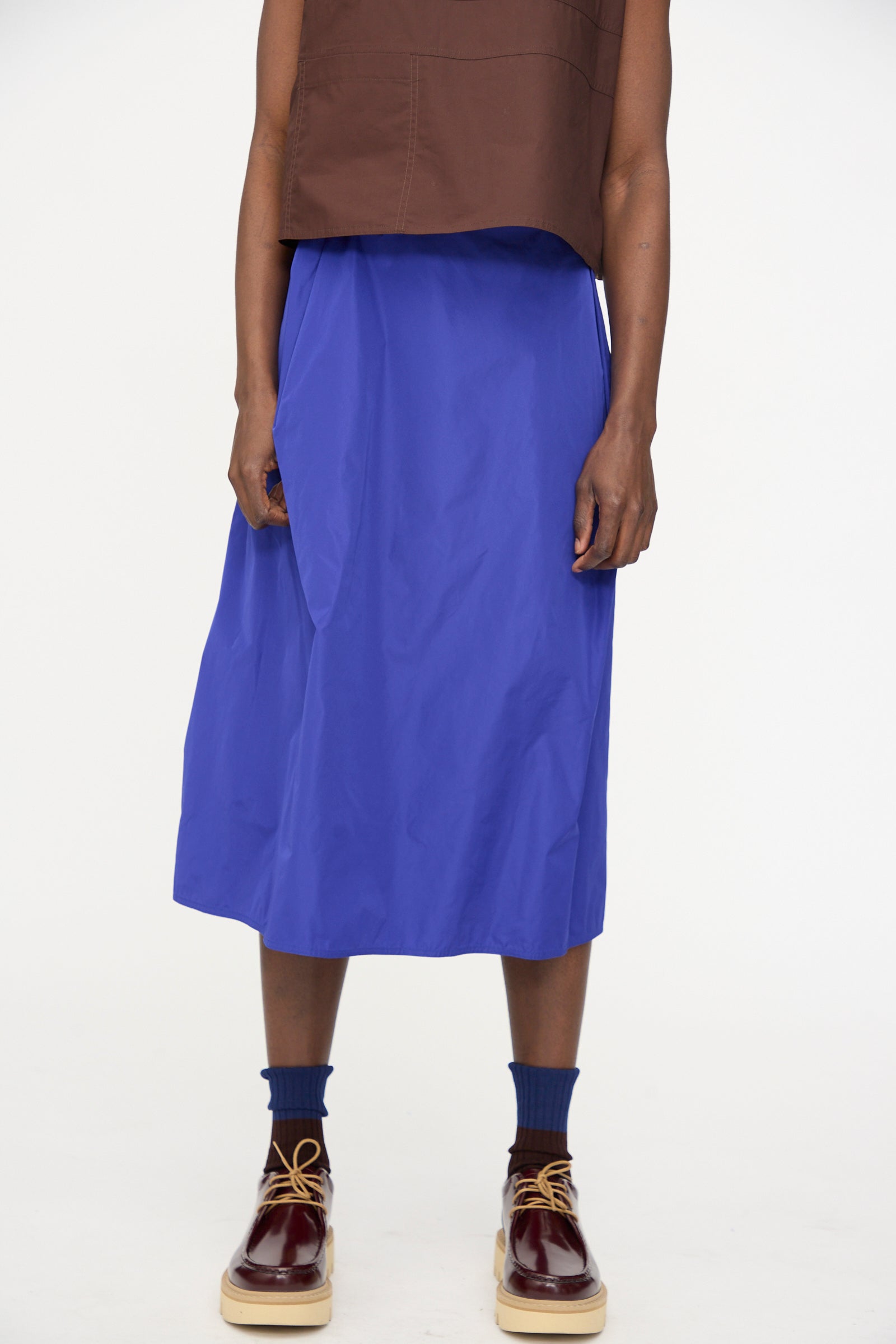 A person wears a brown sleeveless top, the Sofie D’Hoore Taffeta Suvi Skirt in Klein, navy socks, and burgundy platform shoes against a plain white background.