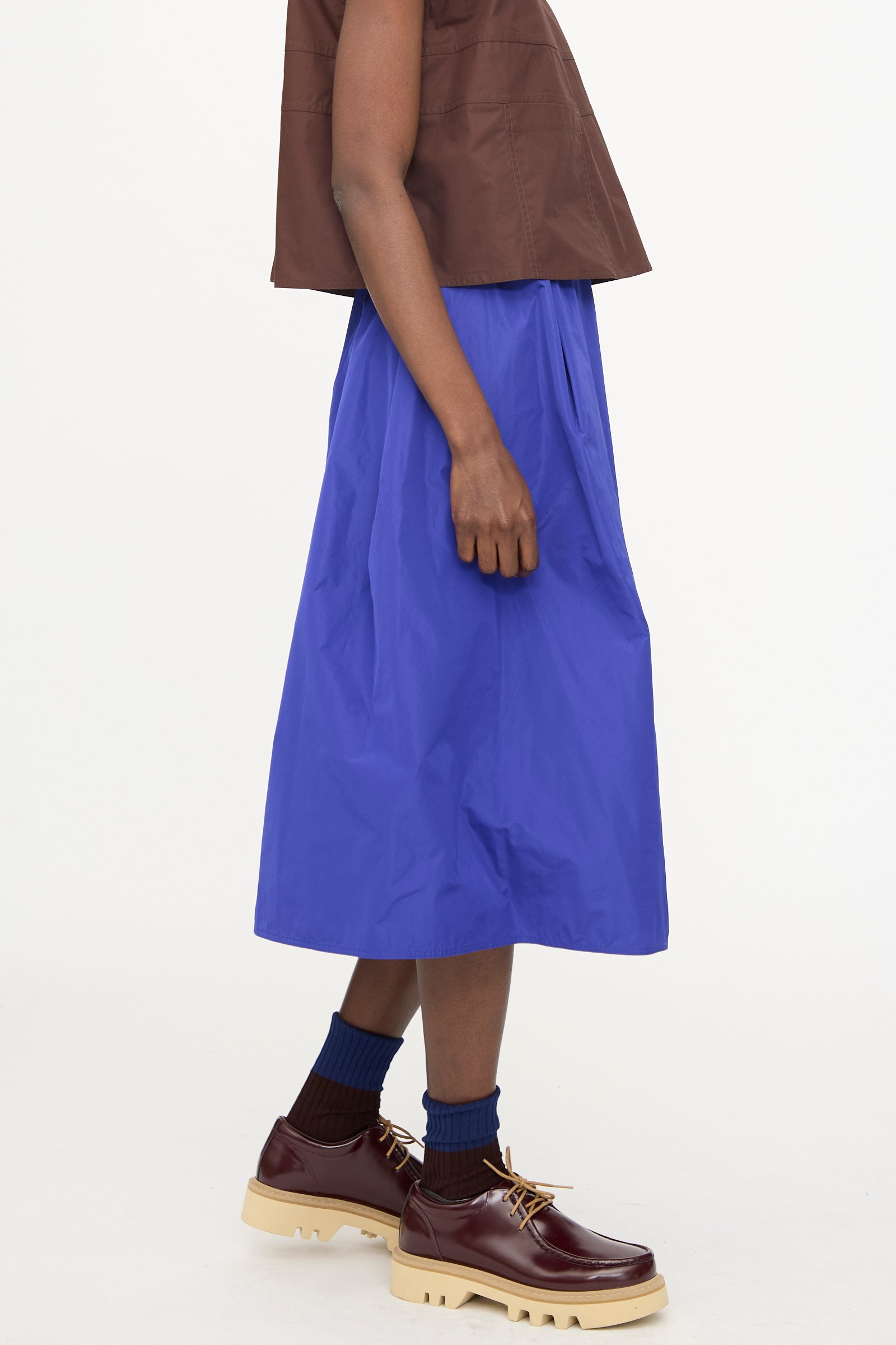 A person stands against a white background wearing the Sofie D'Hoore Taffeta Suvi Skirt in Klein, a brown sleeveless top, blue socks, and brown chunky shoes with tan soles.