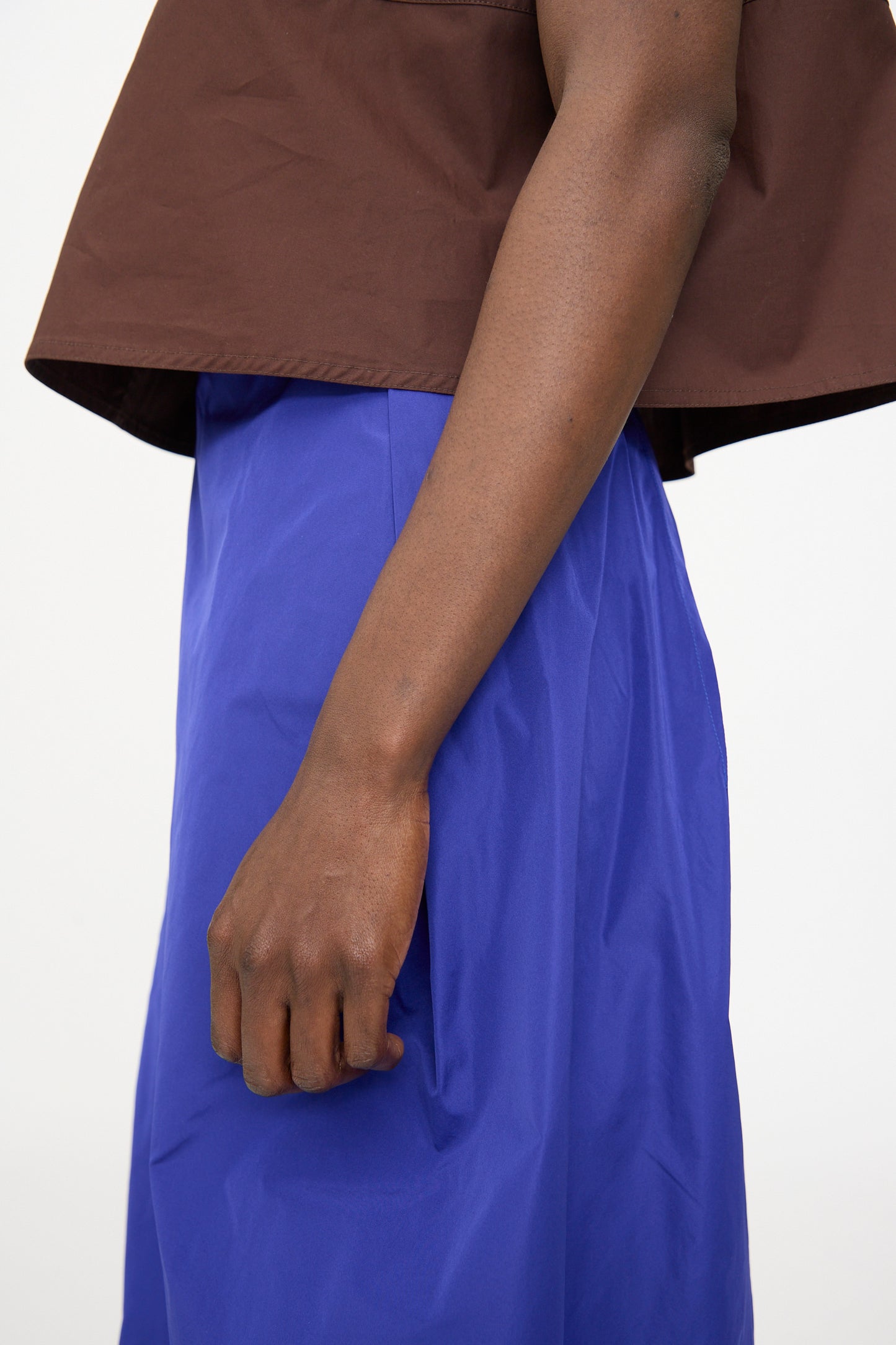 A person stands with their arm at their side, wearing a brown top and the Sofie D'Hoore Taffeta Suvi Skirt in Klein blue against a plain background.