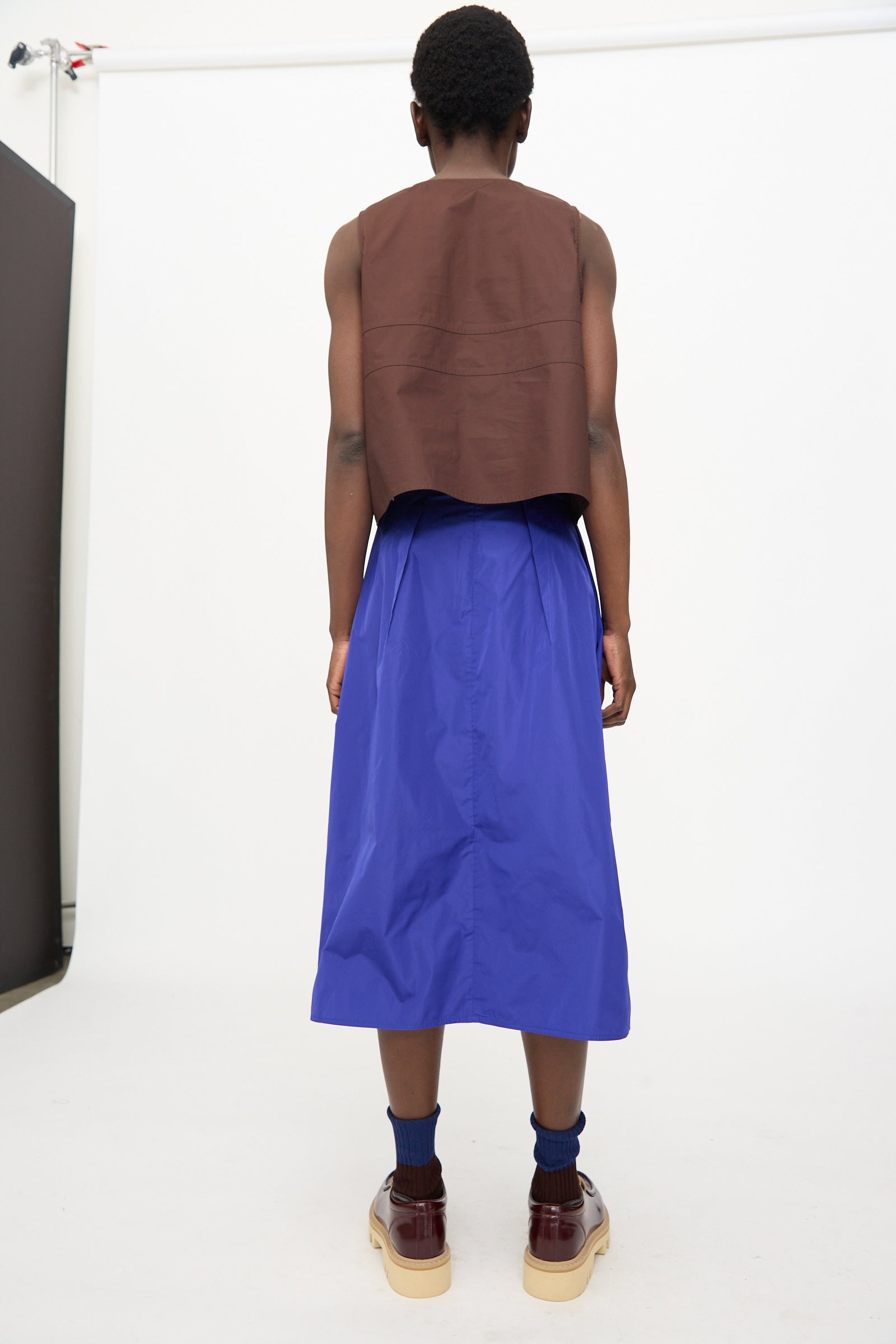 A person stands facing away against a plain white background, wearing a sleeveless brown top, blue socks, burgundy platform shoes, and the Sofie D’Hoore Taffeta Suvi Skirt in Klein.