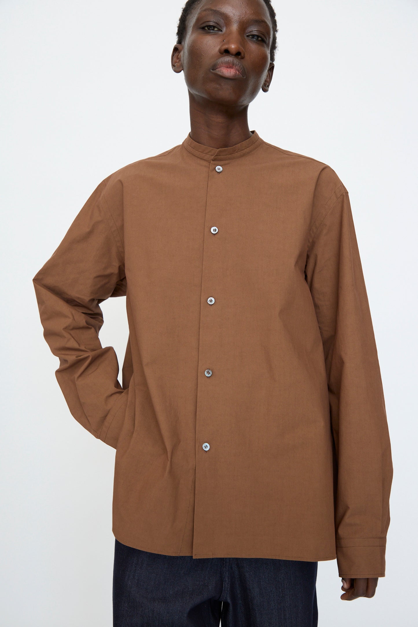 A person wears the Sofie D'Hoore Washed Poplin Braque Shirt in Chestnut, featuring long sleeves and a mandarin collar, standing against a plain light background.