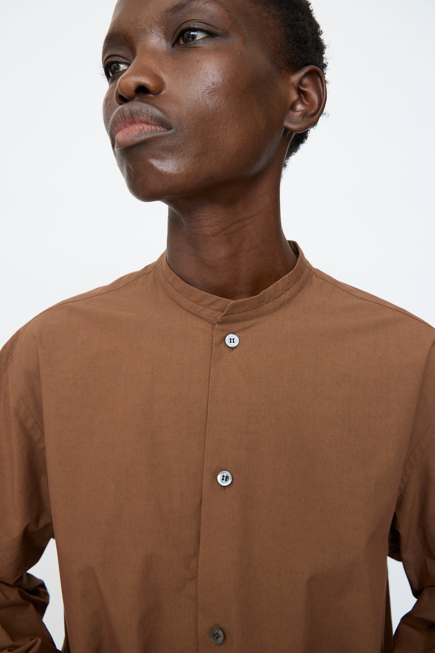 A person wears the Sofie D'Hoore Washed Poplin Braque Shirt in Chestnut, featuring long sleeves and a band collar, standing against a plain white background.