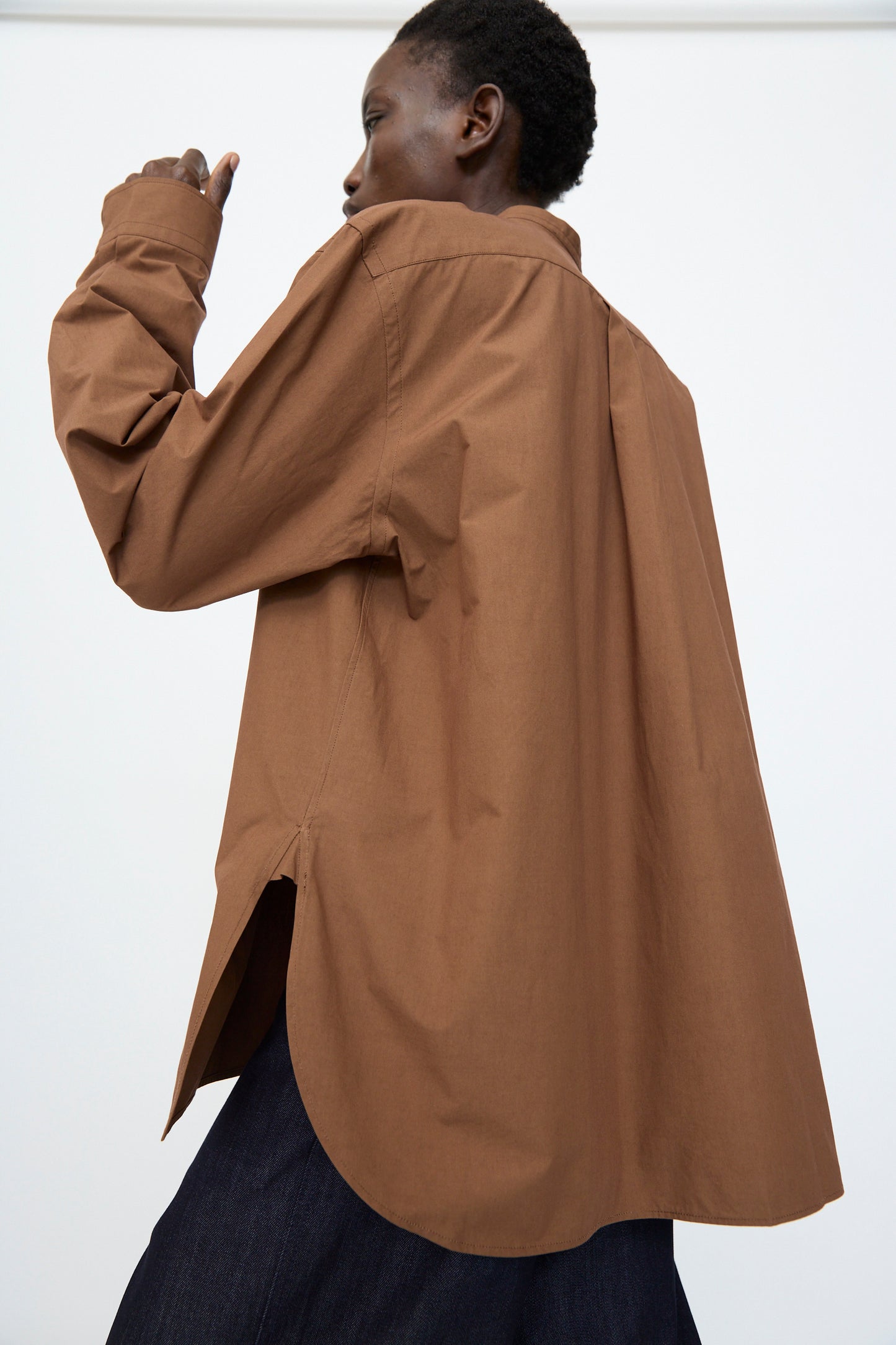 A person models the Sofie D'Hoore Washed Poplin Braque Shirt in Chestnut, a loose-fitting, long-sleeve cotton top with a high-low hem and side slit, paired with dark pants against a plain light background.