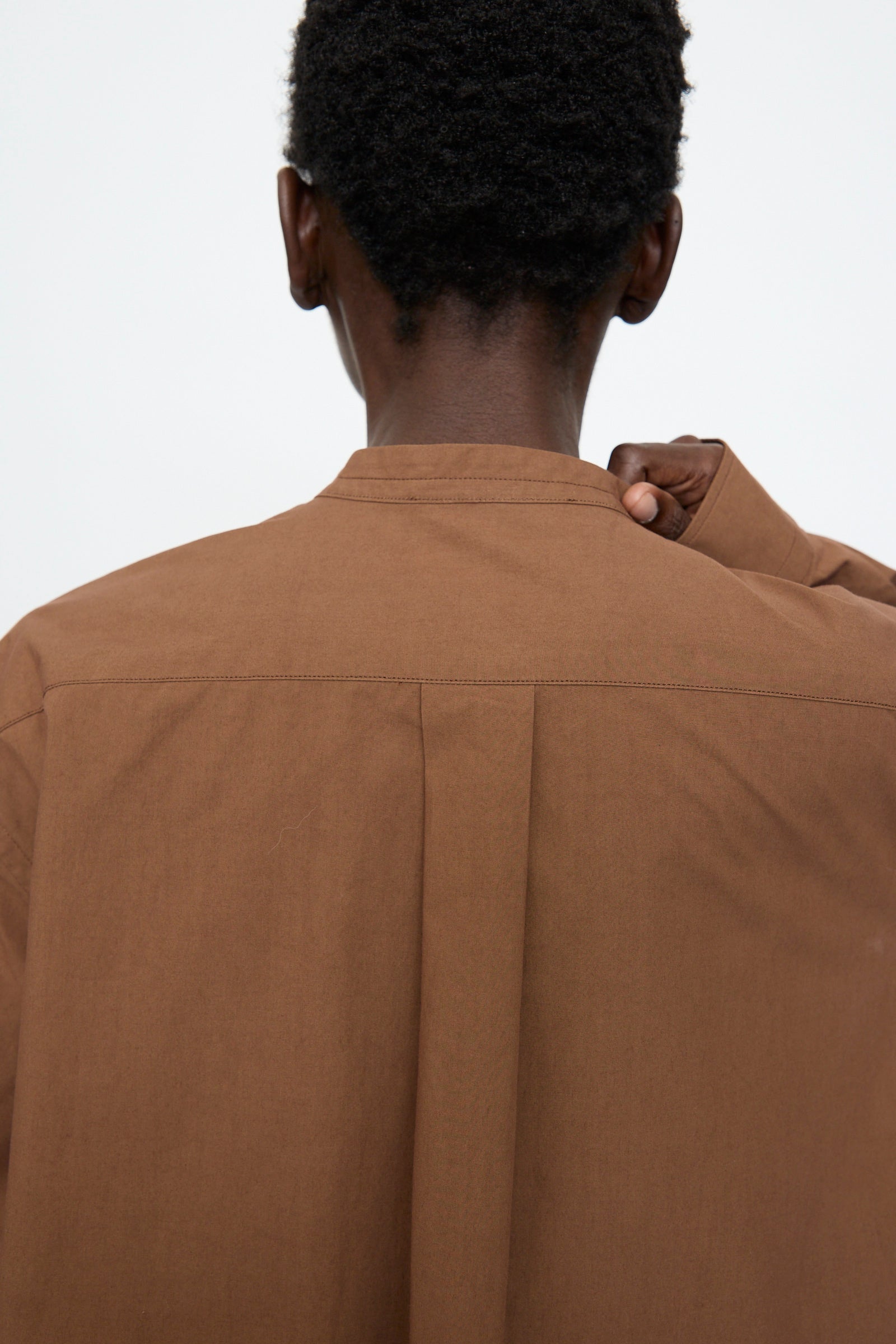 A person seen from behind holds the collar of a Sofie D'Hoore Washed Poplin Braque Shirt in Chestnut, set against a plain light background.