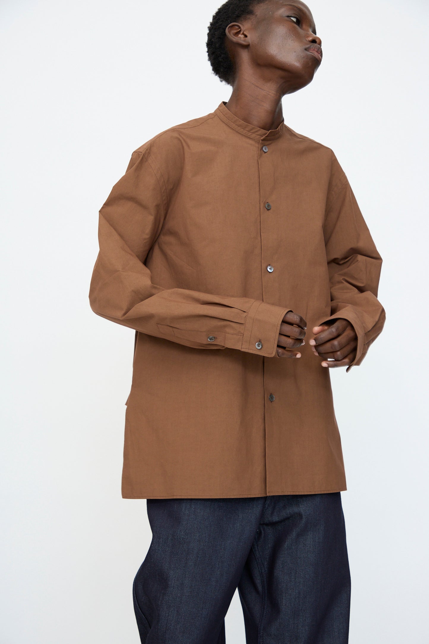 Model in Sofie D'Hoore's Washed Poplin Braque Shirt in Chestnut with long sleeves, paired with dark blue pants, stands against a plain light background.