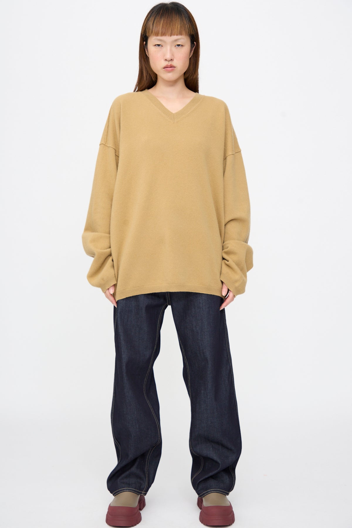 A person wearing a camel-colored Wool Maurice Sweater in an oversized, superfine wool V-neck style with dark denim jeans and platform shoes.
