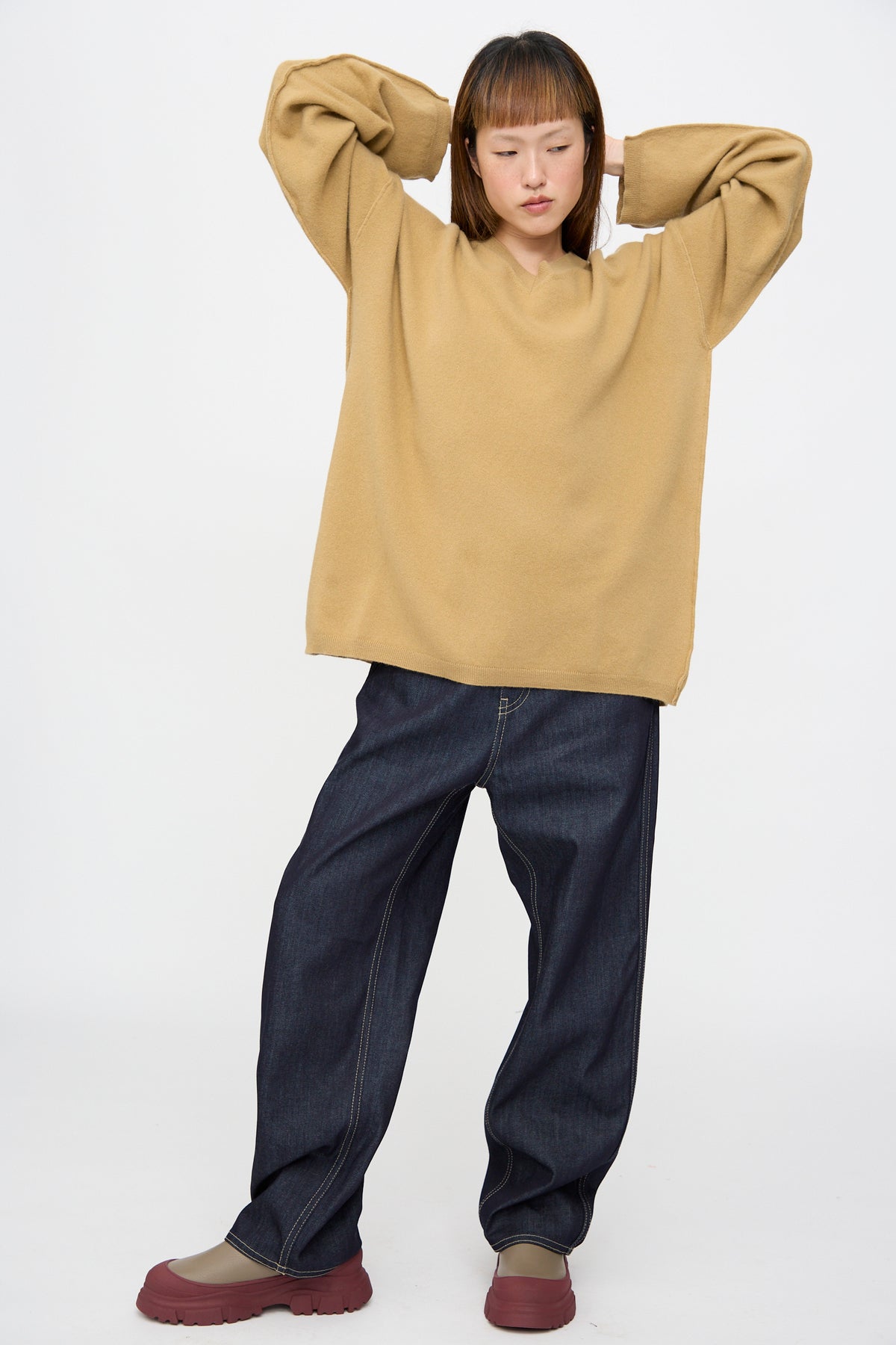 A person wearing a camel Wool Maurice Sweater in a V-neck cut, dark denim wide-leg jeans, and olive green shoes with thick maroon soles.