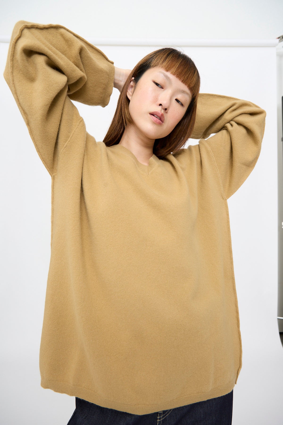 A person wearing the camel Wool Maurice Sweater, an oversized V-neck with wide sleeves, posing with their hands behind their head.