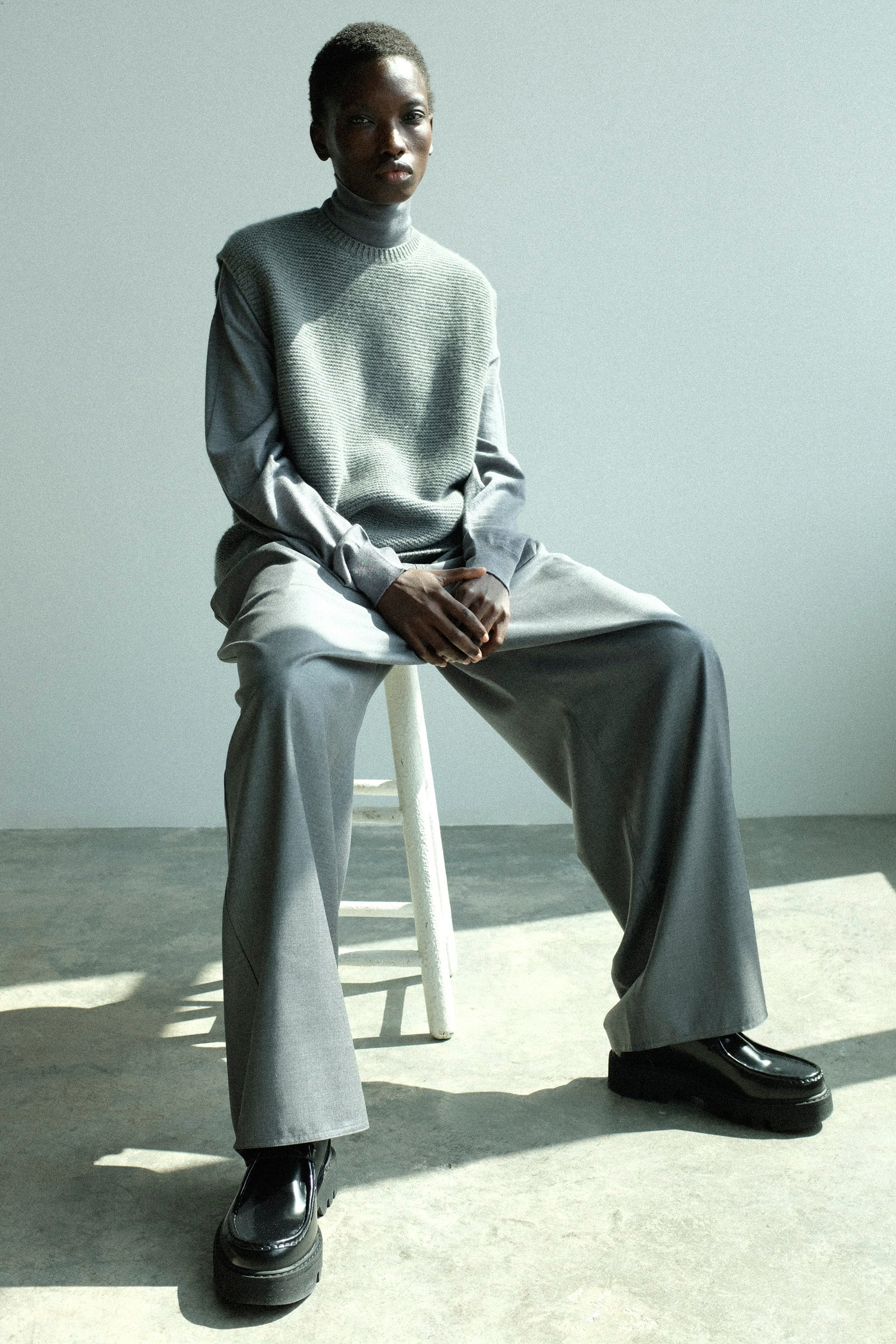 Person wearing a Sofie D'Hoore's Cashmere Matta Sweater in Mid Grey Melange with the Merino Maffei Sweater in Steel Melange, and Superfine Wool Pasch Pants in Mid Grey Melange, sitting on a white stool.