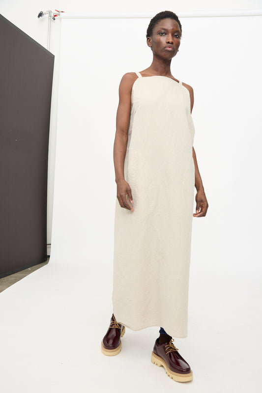 A person stands on a white backdrop wearing the Sofie D'Hoore Linen Cotton Dele Dress in Natural, styled with brown lace-up shoes featuring thick soles.