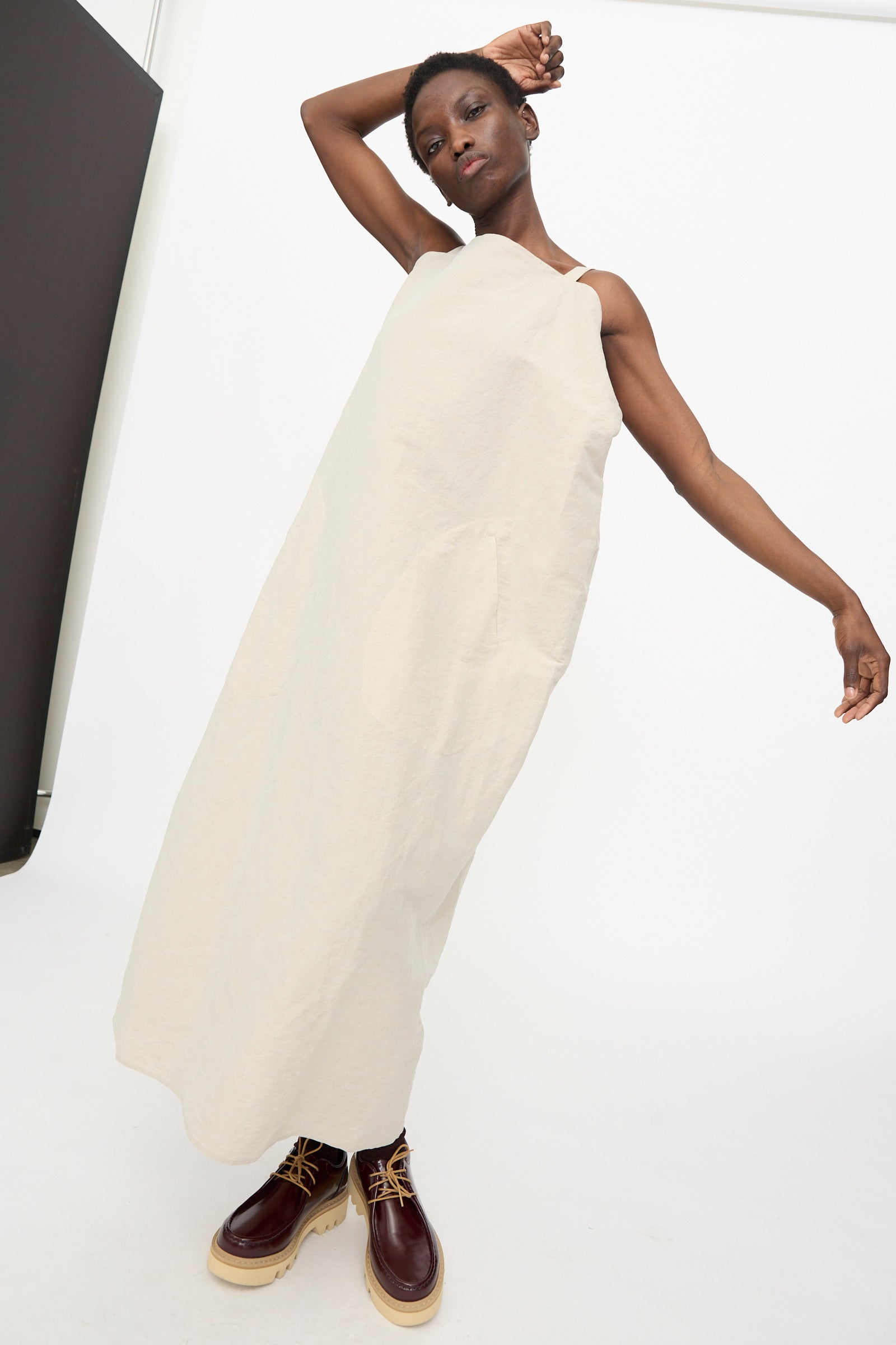 A person stands in a minimalist studio wearing the Sofie D'Hoore Linen Cotton Dele Dress in Natural, paired with brown lace-up shoes, one arm raised and the other extended outward.