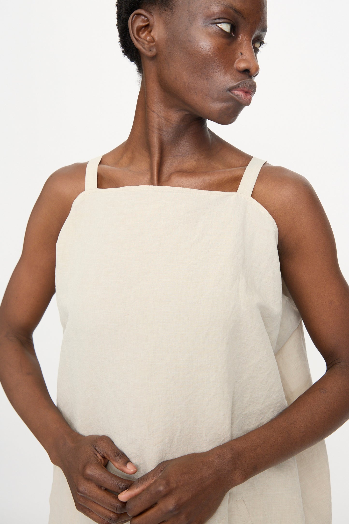 A person models the Sofie D'Hoore Linen Cotton Dele Dress in Natural, standing against a white background and looking to the side with hands clasped.
