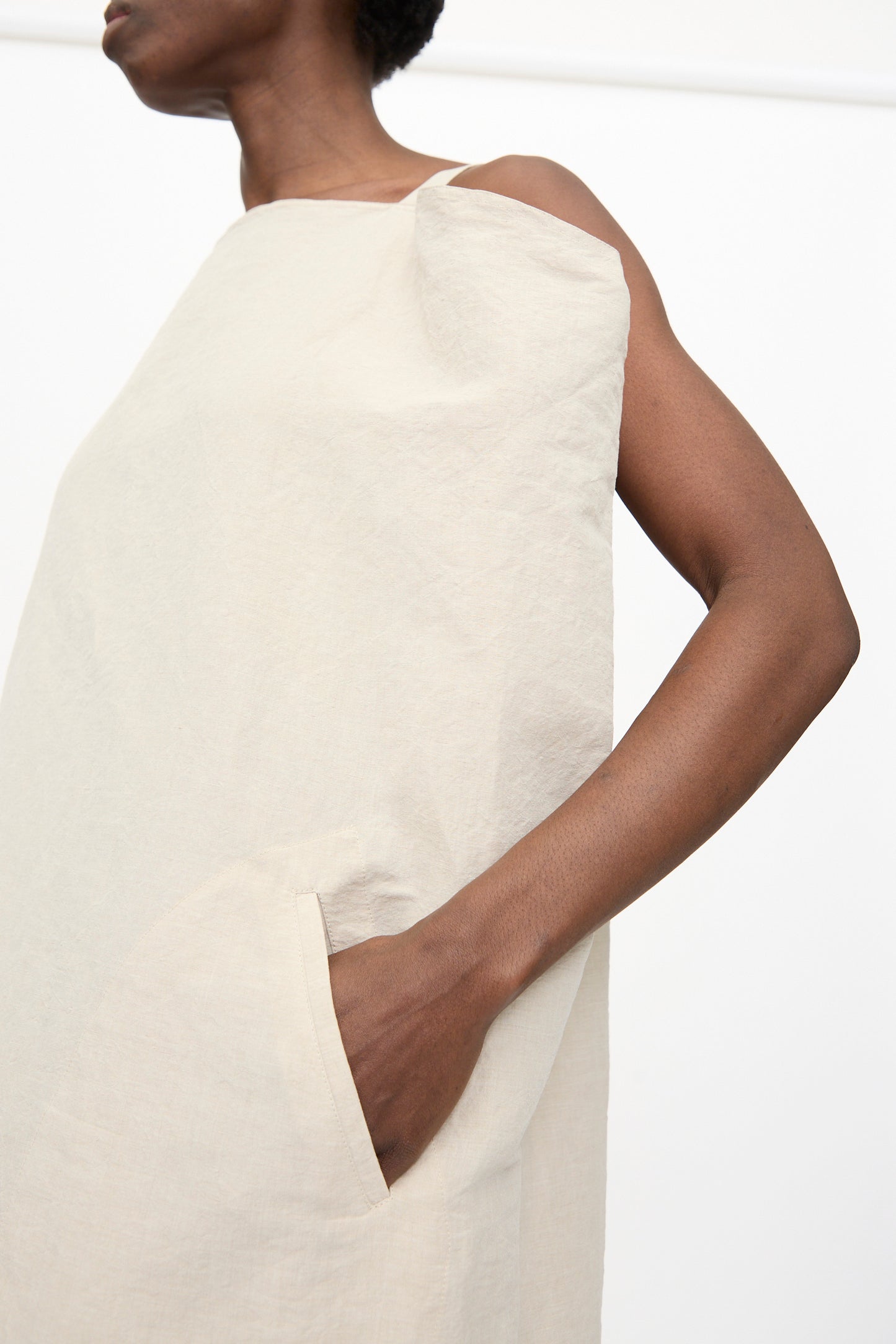 A person wears the Sofie D'Hoore Linen Cotton Dele Dress in Natural, a sleeveless maxi dress with side pockets, standing against a plain white background.