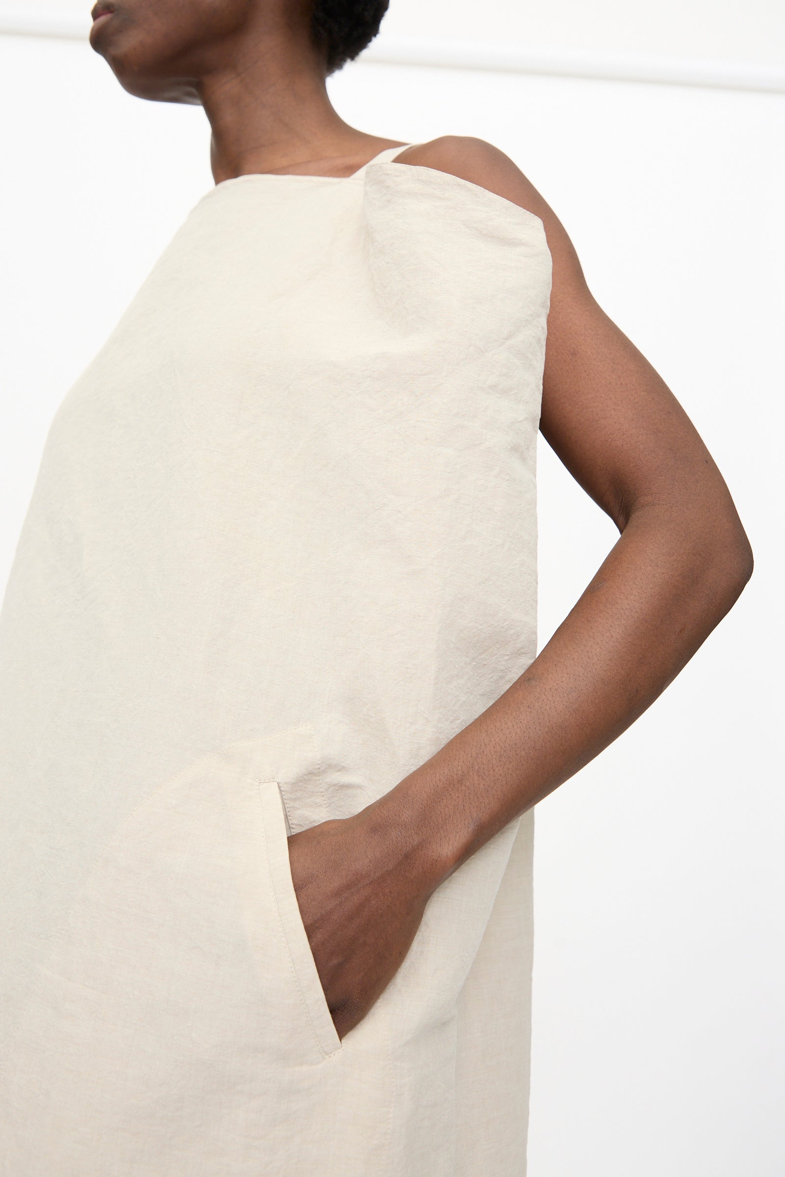 A person wears the Sofie D'Hoore Linen Cotton Dele Dress in Natural, a sleeveless maxi dress with side pockets, standing against a plain white background.
