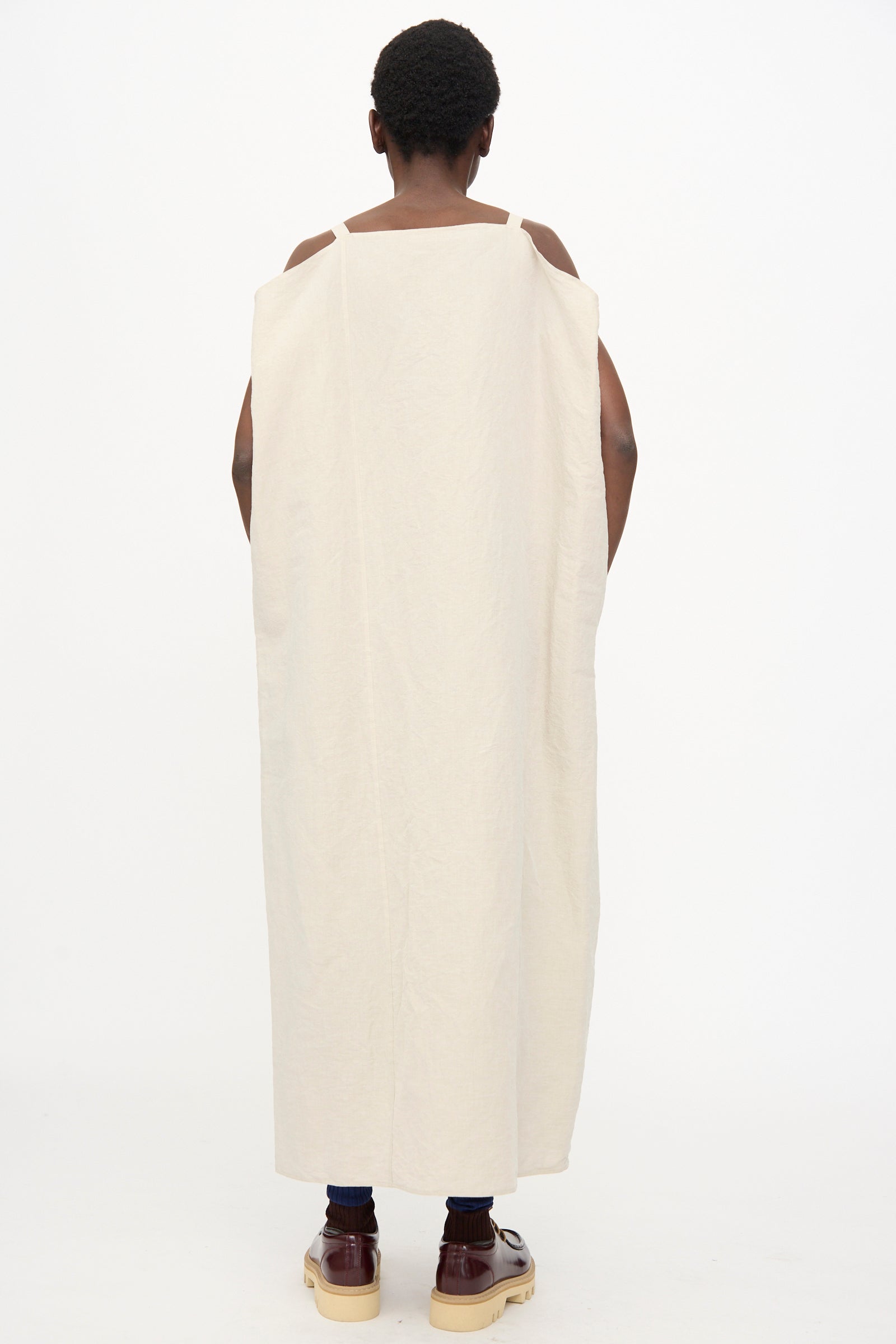 A person stands facing away, wearing the Sofie D'Hoore Linen Cotton Dele Dress in Natural—a long, loose, sleeveless off-white maxi dress with a square neckline—paired with brown shoes against a plain white background.