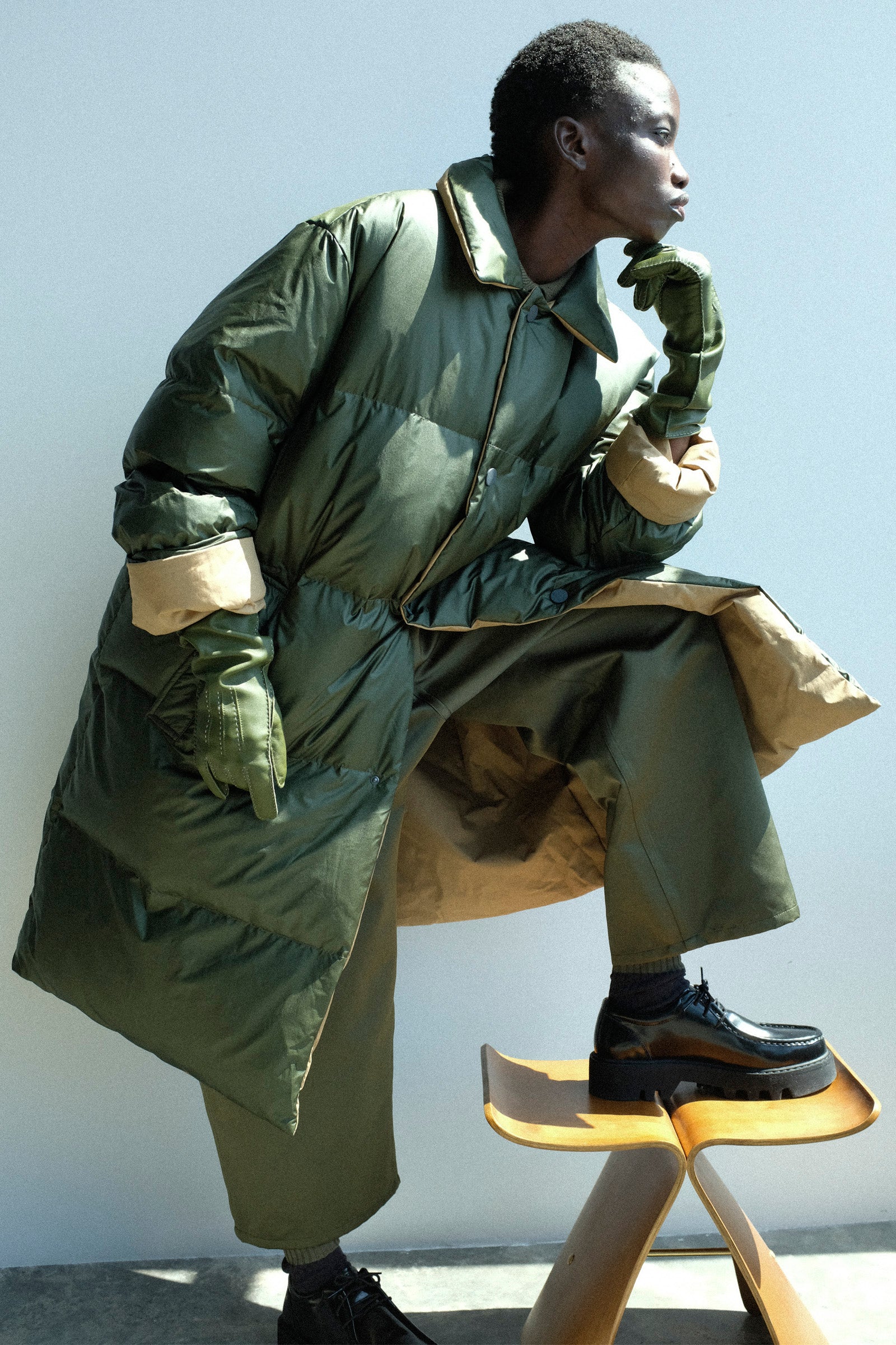 Person wearing a Sofie D'Hoore's Reversible Otis Down Coat in Mushroom and Khaki, Leather Vasarely Gloves in Moss, Satin Polke Pant in Laurel, and the Leather Falcone Wallabee Shoe in Black against a light background.