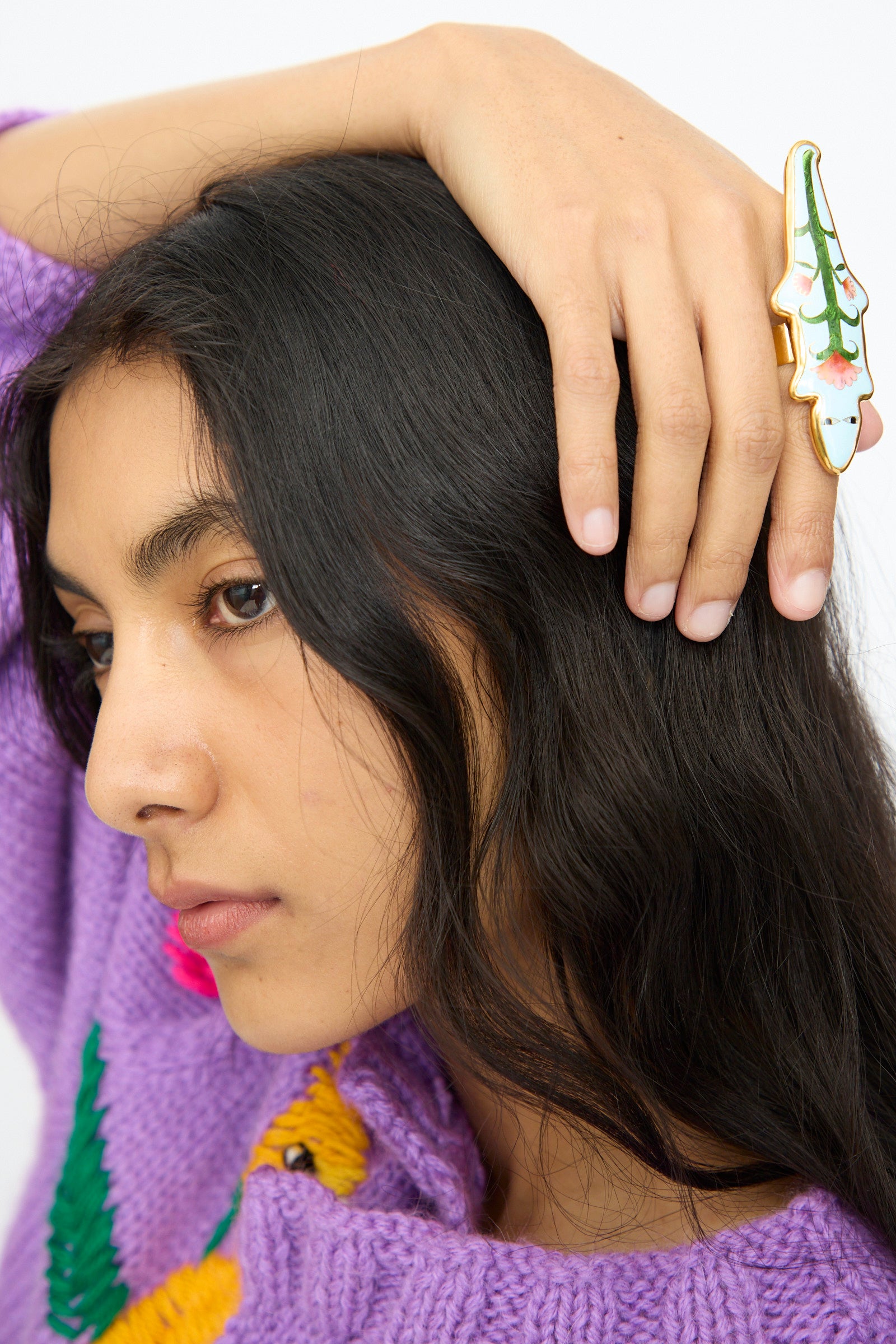 A person with long dark hair rests their hand on their head, wearing the Sofio Gongli Alligator Ring in Blue. They are dressed in a purple sweater with a yellow design.