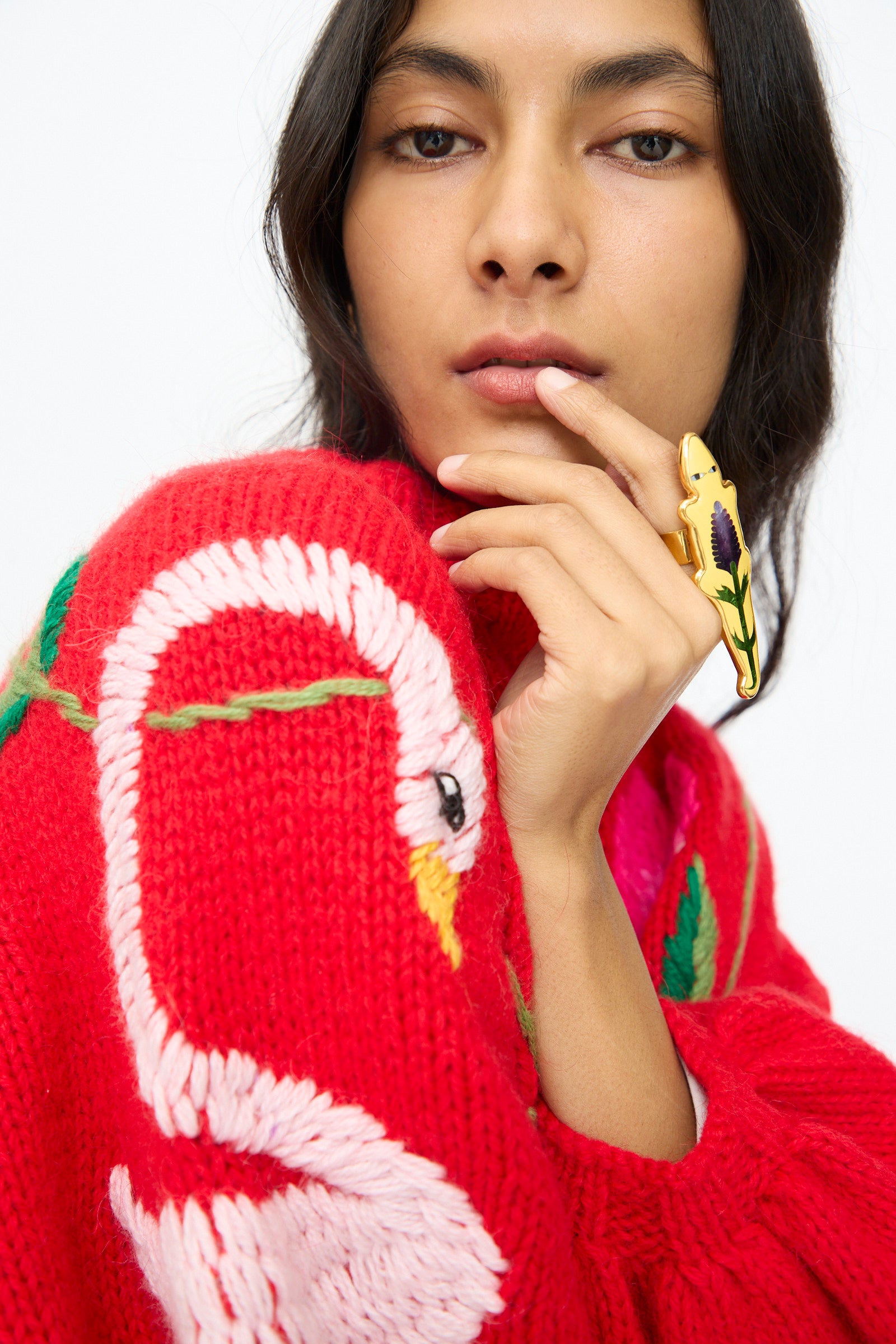 A woman wearing a red knit sweater with a white swan design poses with her hand near her face, showcasing the Sofio Gongli Alligator Ring in Yellow.