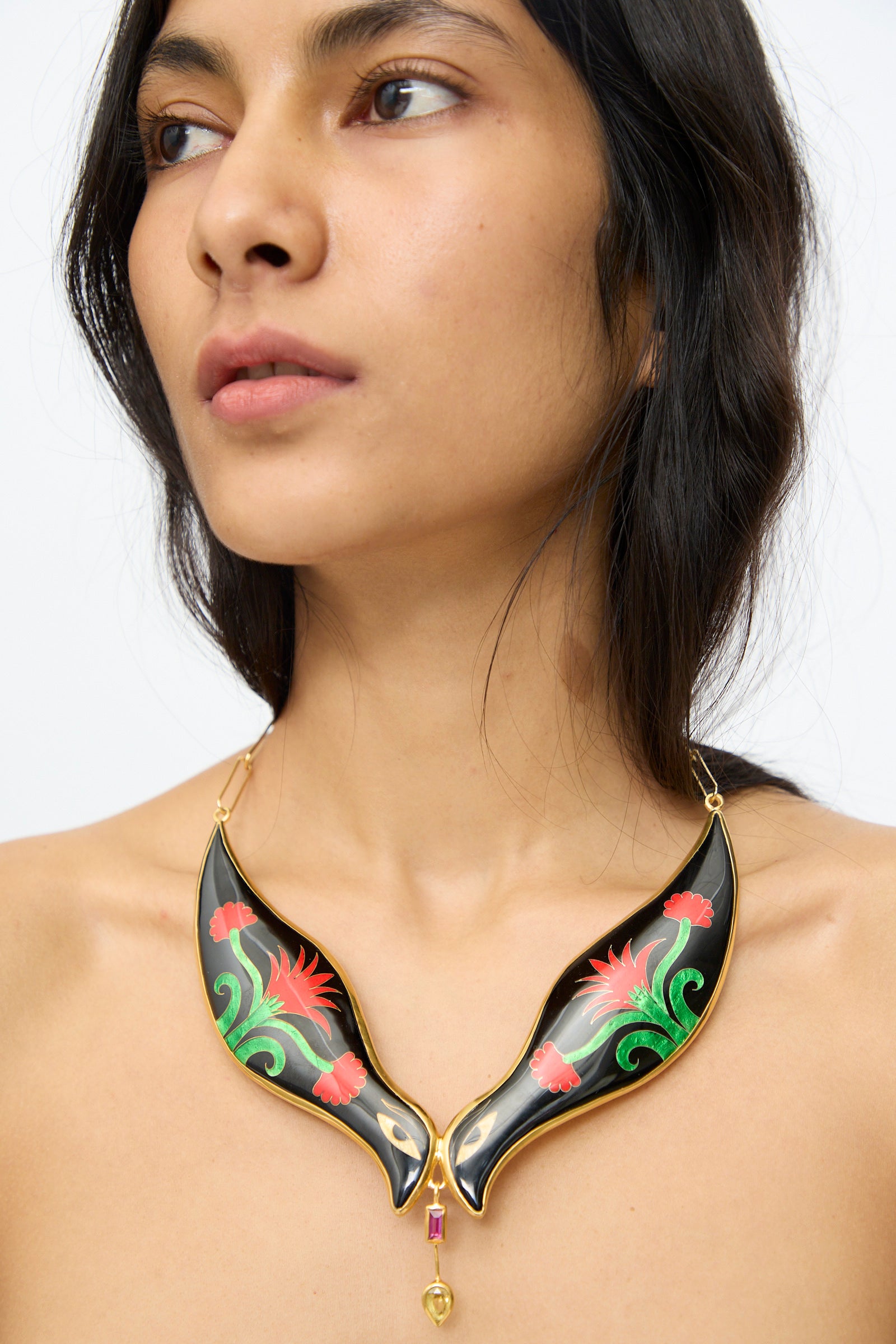A person wearing the Sofio Gongli Bird Necklace in Black and Red Flower, featuring a leaf-shaped pendant with red and green floral designs, gazes slightly to the side.
