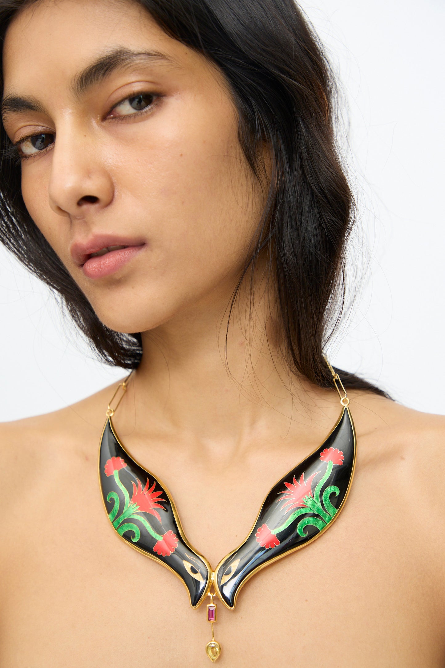 A woman with long dark hair wears the Sofio Gongli Bird Necklace in Black and Red Flower, a statement piece featuring wing-shaped cloisonné sections adorned with red and green floral designs.