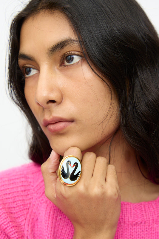A person with long dark hair and a pink sweater, holding their chin, showcases the Sofio Gongli Black Swan Ring—an adjustable gold-plated piece featuring a striking black swan design.