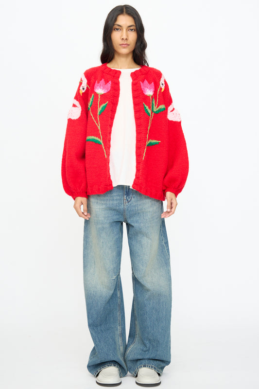 A woman stands facing forward wearing the Sofio Gongli Cardigan in Red with Flowers and Swans over a white shirt, paired with wide-leg blue jeans and white shoes against a plain white background.