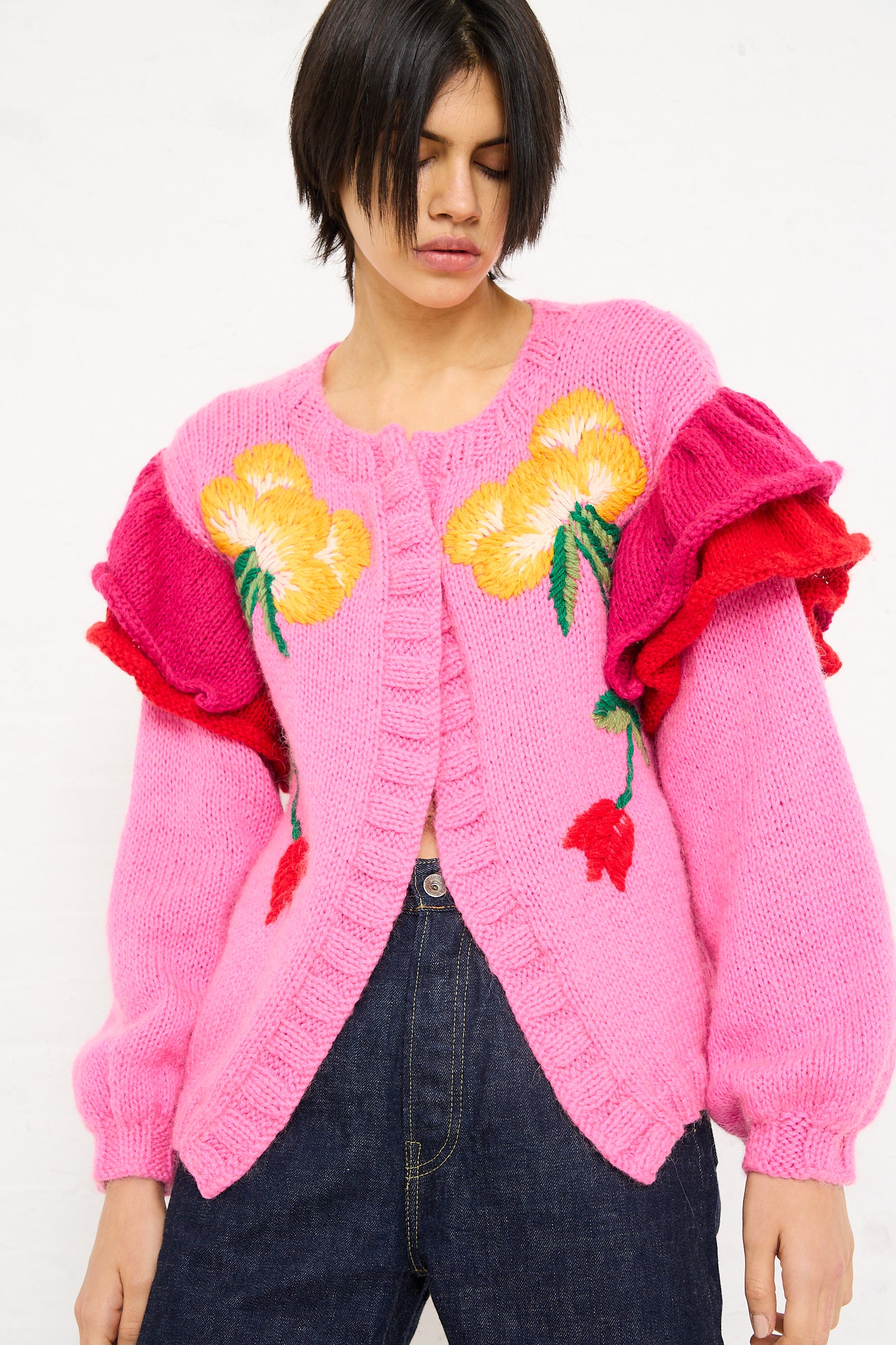 Woman in a Sofio Gongli pink, hand-knit open cardigan with large yellow floral embroidery and voluminous sleeves, designed by a Georgian knitwear designer, standing against a white background.