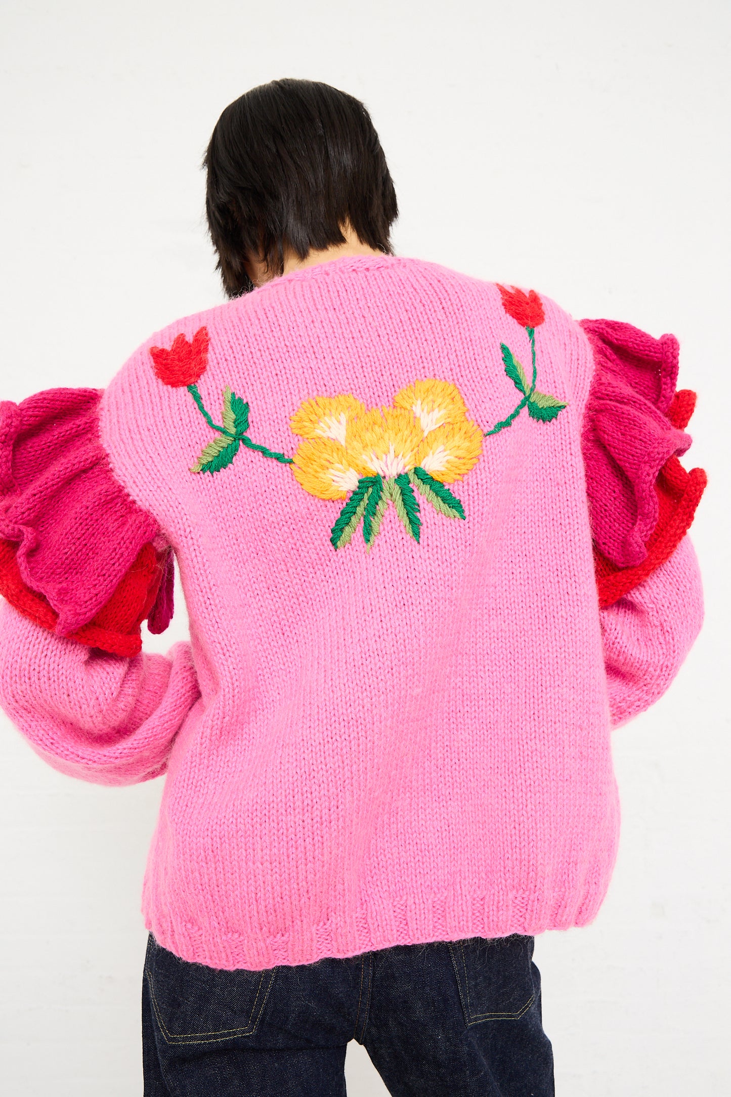 A person from behind wearing a Sofio Gongli pink hand-knit open cardigan with a large yellow flower and green leaves embroidered on it.