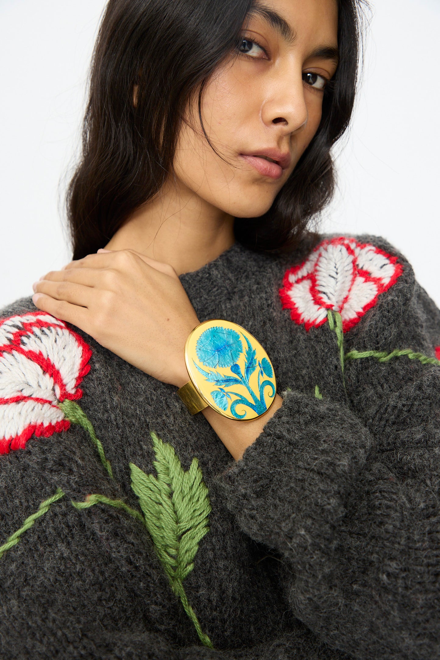 A woman in a grey sweater with red and white floral embroidery highlights the Sofio Gongli Flower Bracelet in Blue, featuring a striking blue enamel design on a 14K gold-plated cuff.