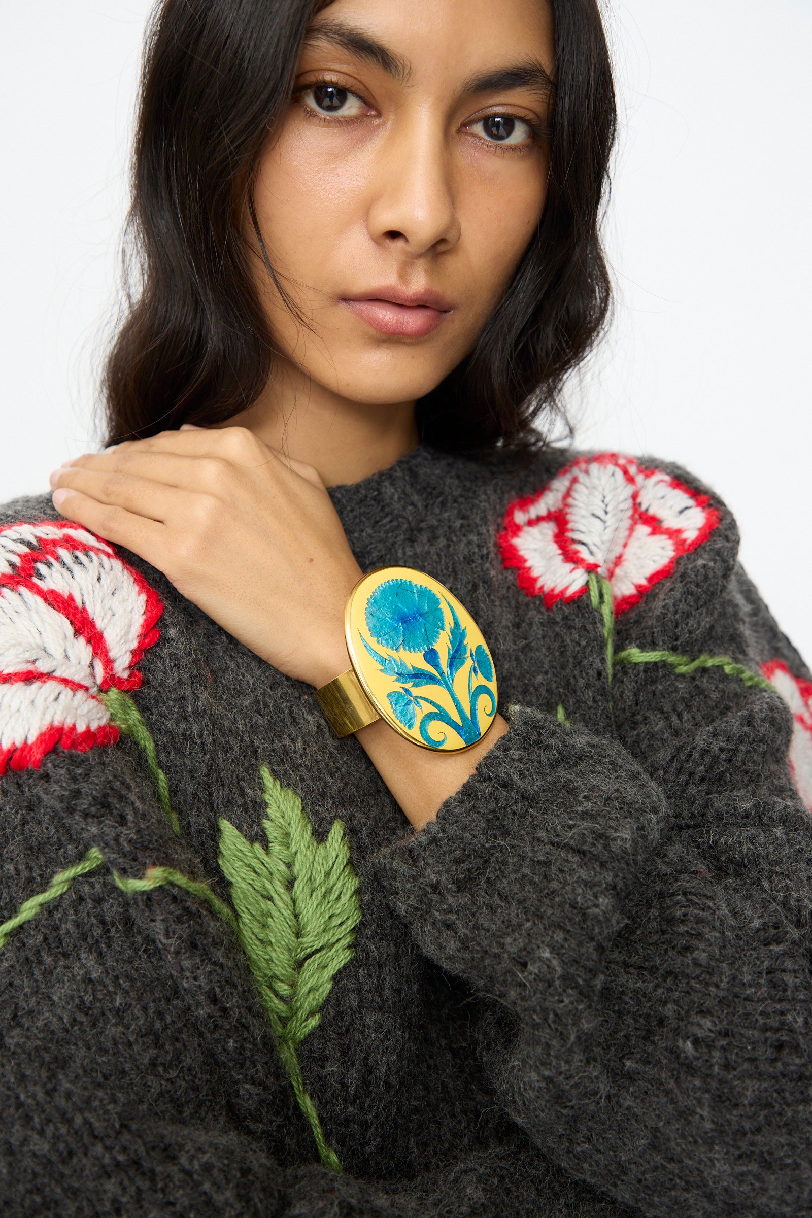 Someone wearing a dark gray sweater with red and white flower embroidery displays the Sofio Gongli Flower Bracelet in Blue, a 14K gold plated cuff featuring a blue floral design, on their wrist.