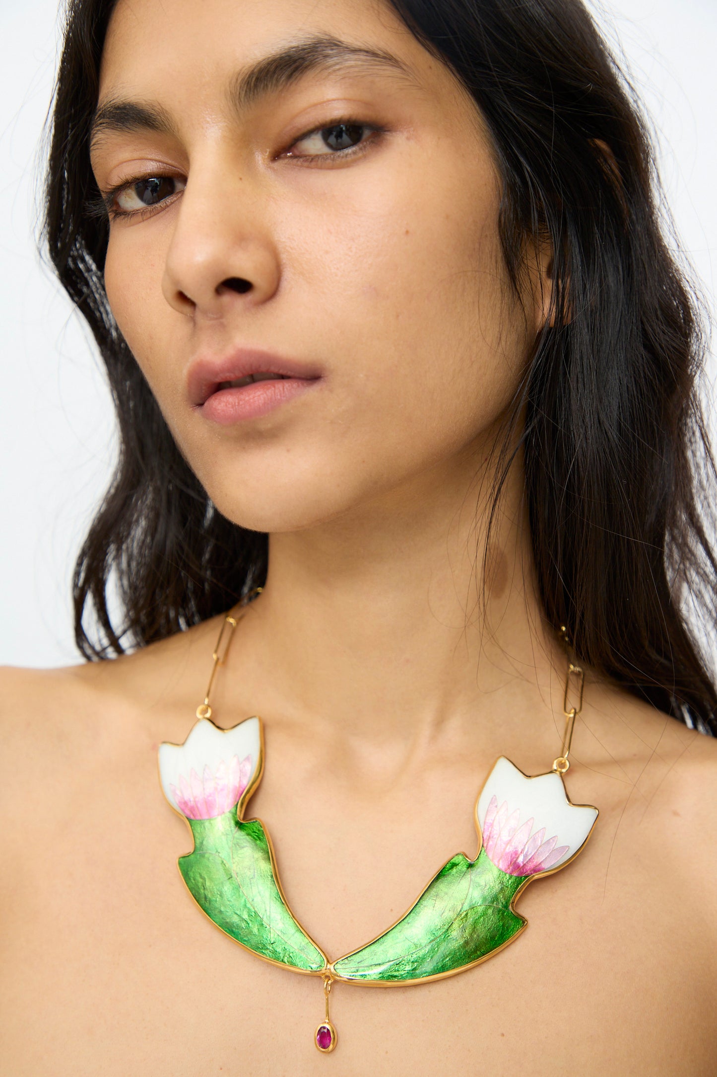 A person with long dark hair wears the Sofio Gongli Flower Necklace, a 14k gold plated piece featuring green and white lotus flowers and a pink gemstone pendant.