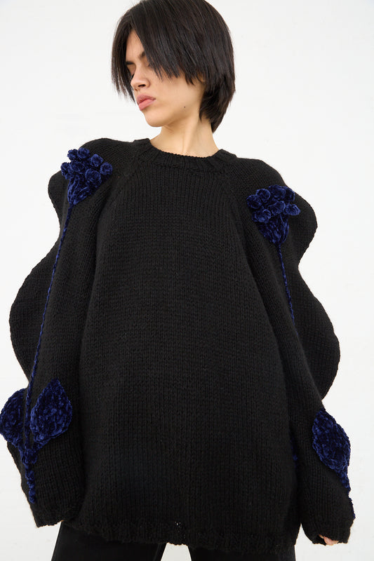 A person with a short haircut is wearing an oversized black hand-knit Sofio Gongli Flower Sweater in Black with blue floral embellishments on the sleeves.