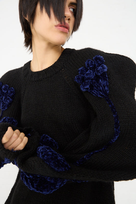 A person with short hair, wearing a Sofio Gongli Flower Sweater in Black with distinctive blue textured details on the sleeves, posing against a light background.