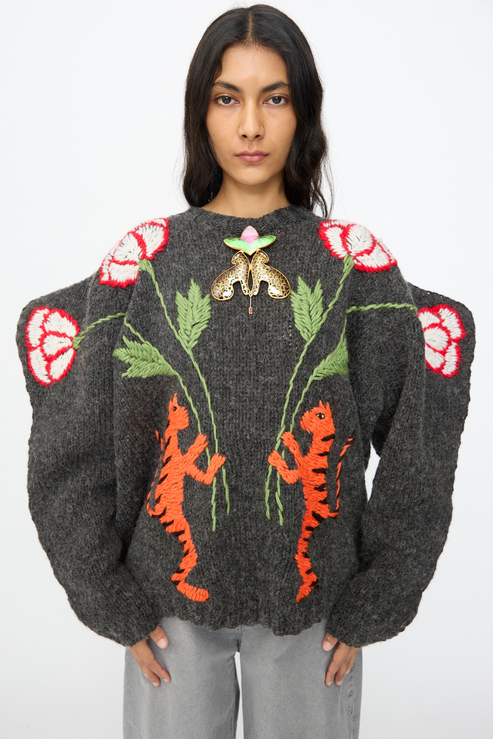 A woman wearing a gray chunky knit sweater with red and white flowers, orange leopard designs, and the Sofio Gongli Leopard and Flower Pendant stands against a plain background.