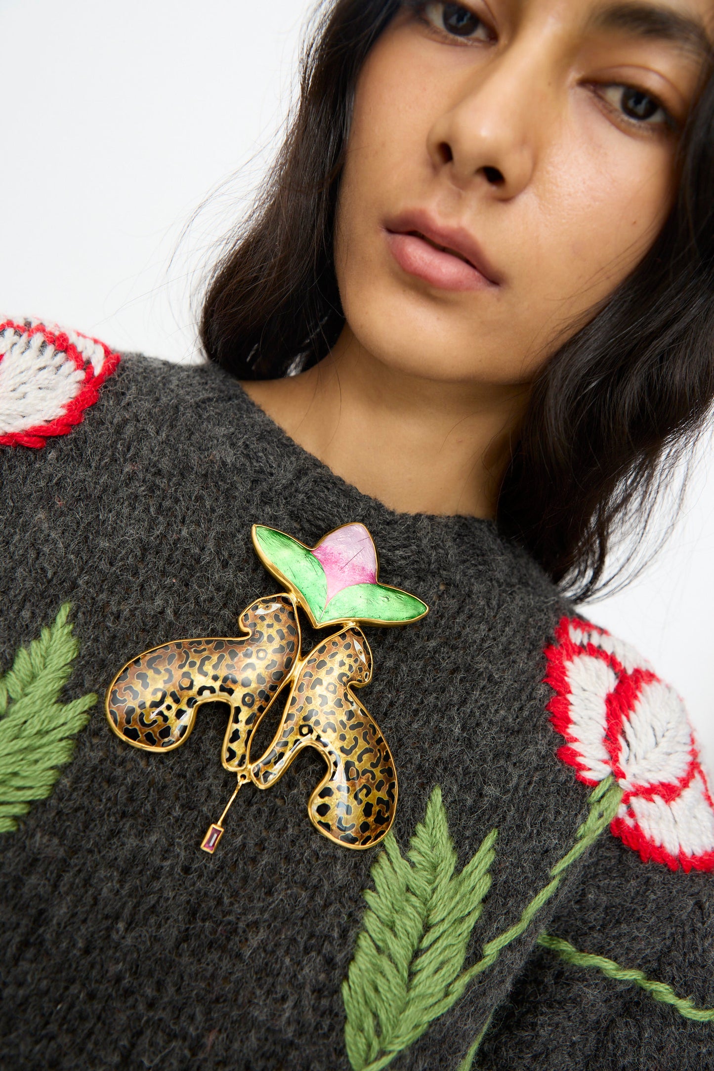 A woman wears a dark knitted sweater with red and white flower patterns, accessorized with the Sofio Gongli Leopard and Flower Pendant: a cloisonné piece featuring leopard print petals and a pink-green center.