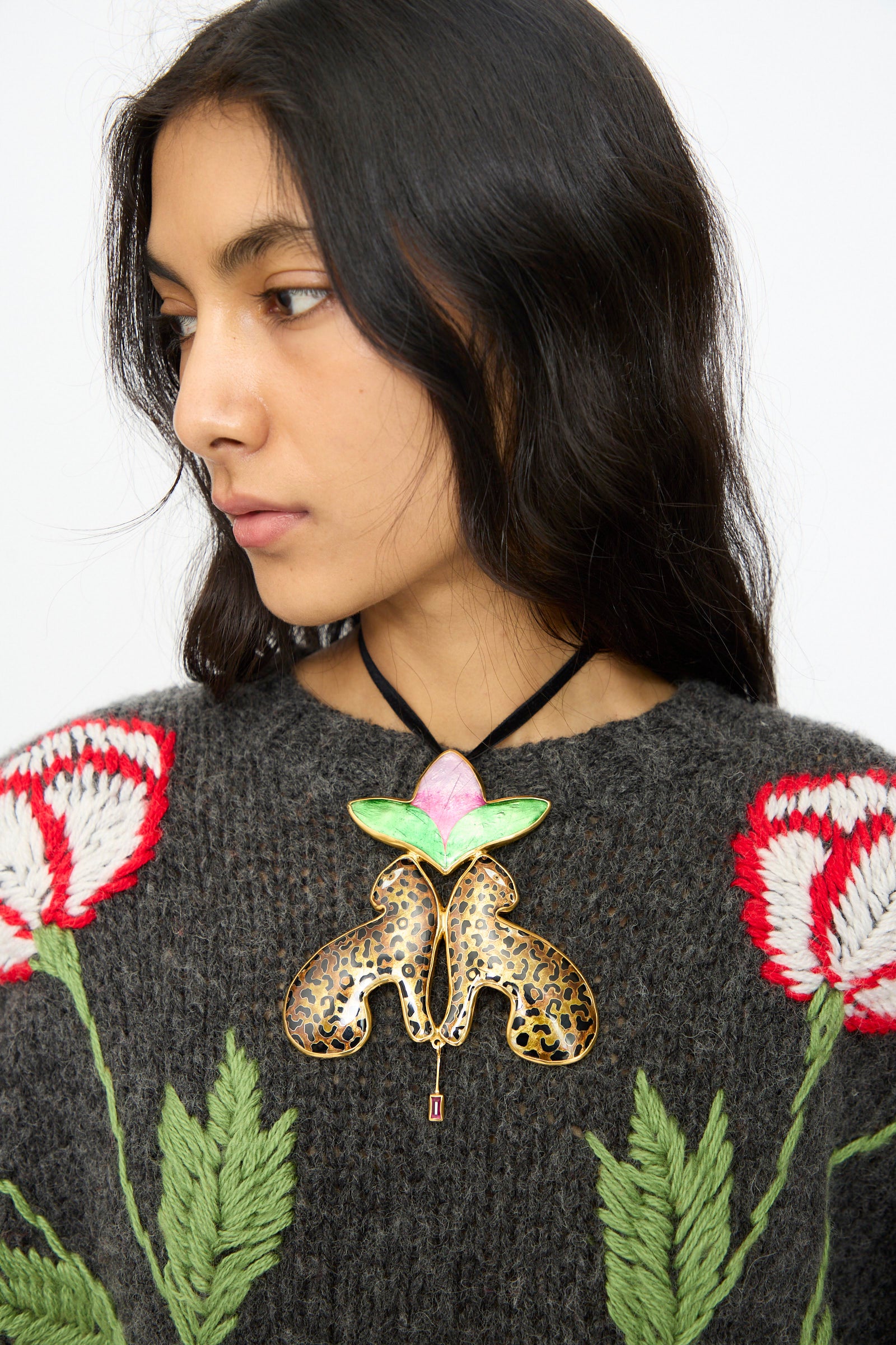 A woman with long dark hair wears a gray sweater with red and white flowers, accessorized by the Sofio Gongli Leopard and Flower Pendant—14K gold plated enamel, butterfly-shaped, with green, pink, and gold details.