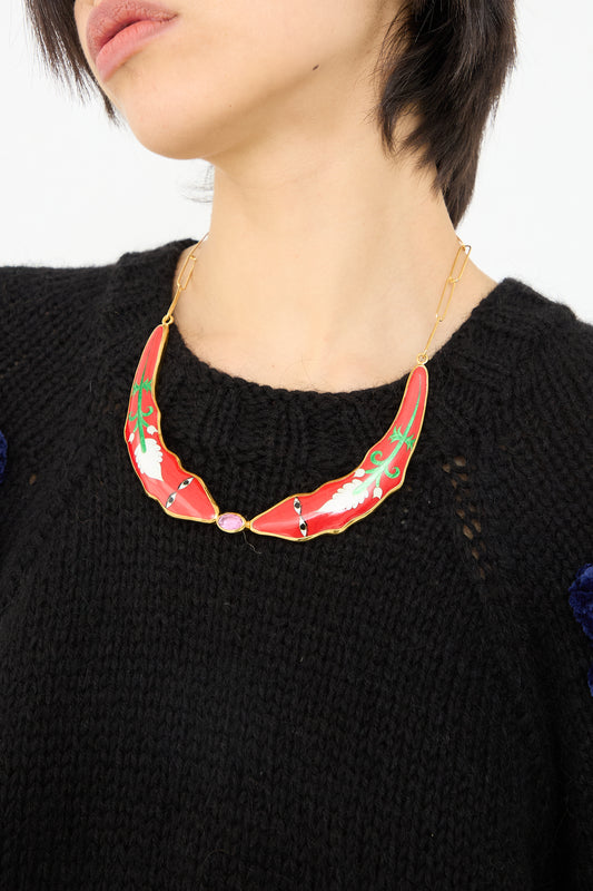 A close-up image of a person wearing a black sweater and an ornate Sofio Gongli cloisonné necklace with gold chain and red, green, and purple enamel accents.