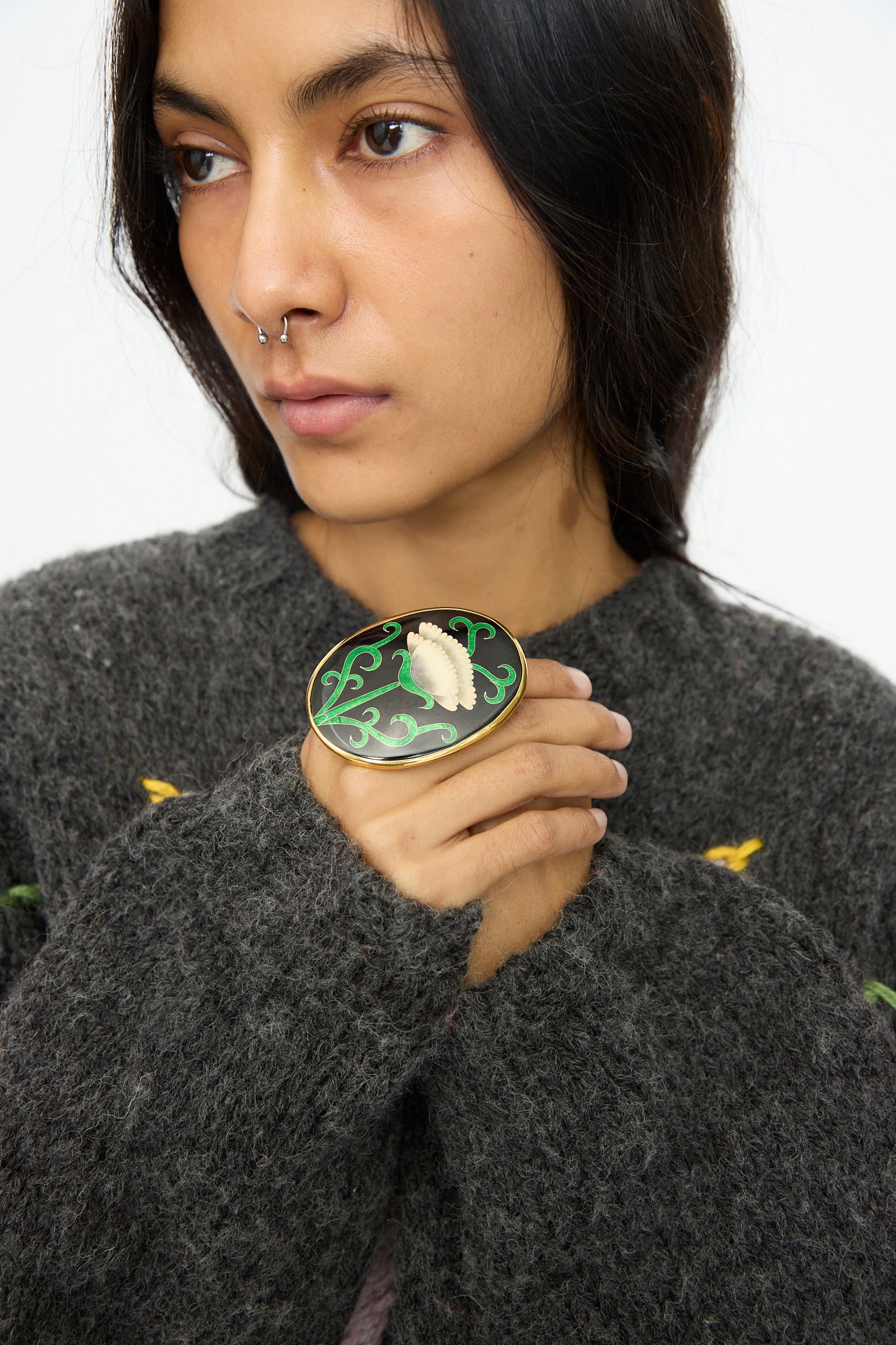 Person with long dark hair and a nose ring wears a textured gray sweater, holding the Sofio Gongli Oval Ring in Black with White Flower.
