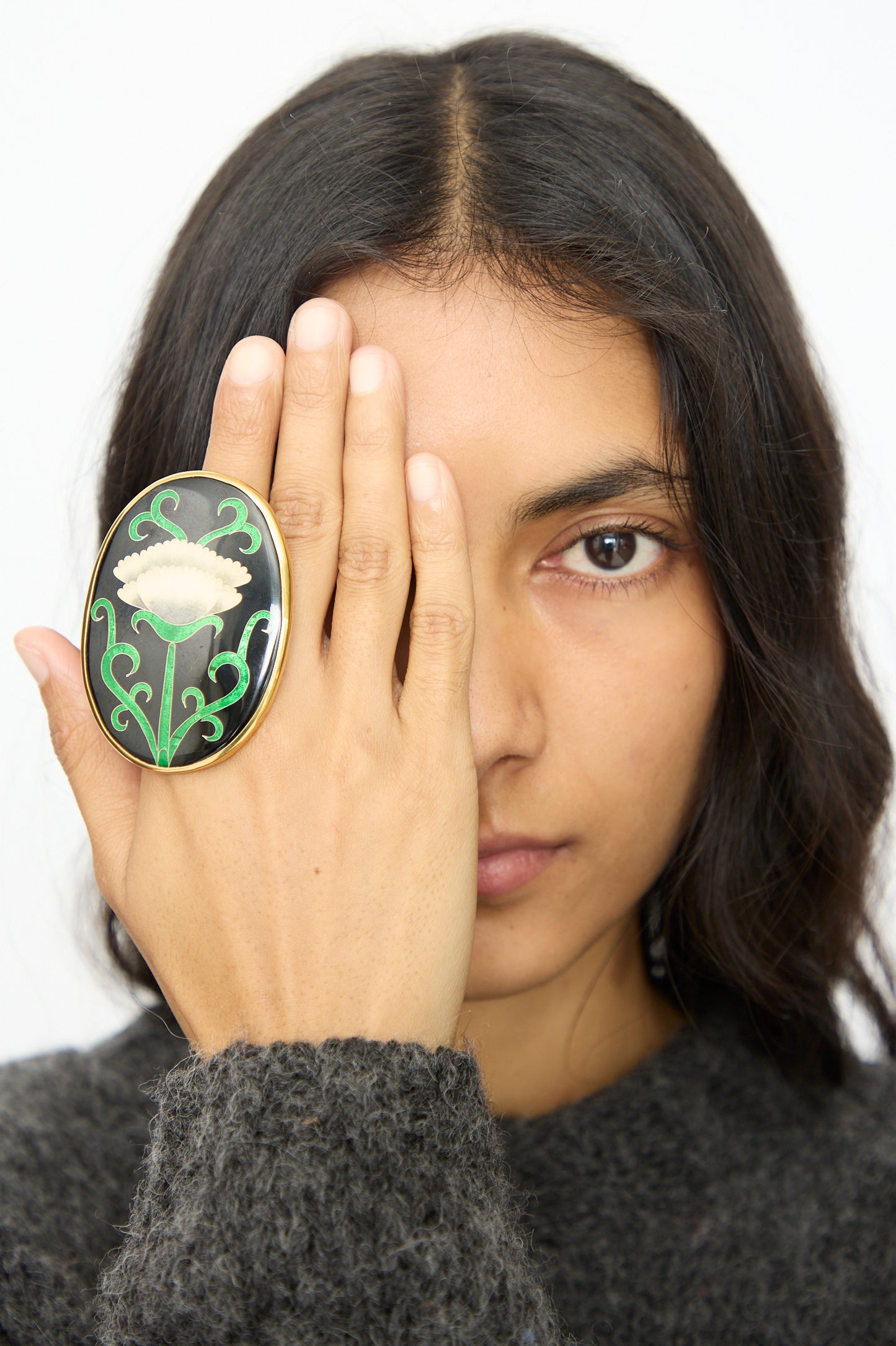 A person with long dark hair covers one eye with their hand, wearing the Sofio Gongli Oval Ring in Black with White Flower.