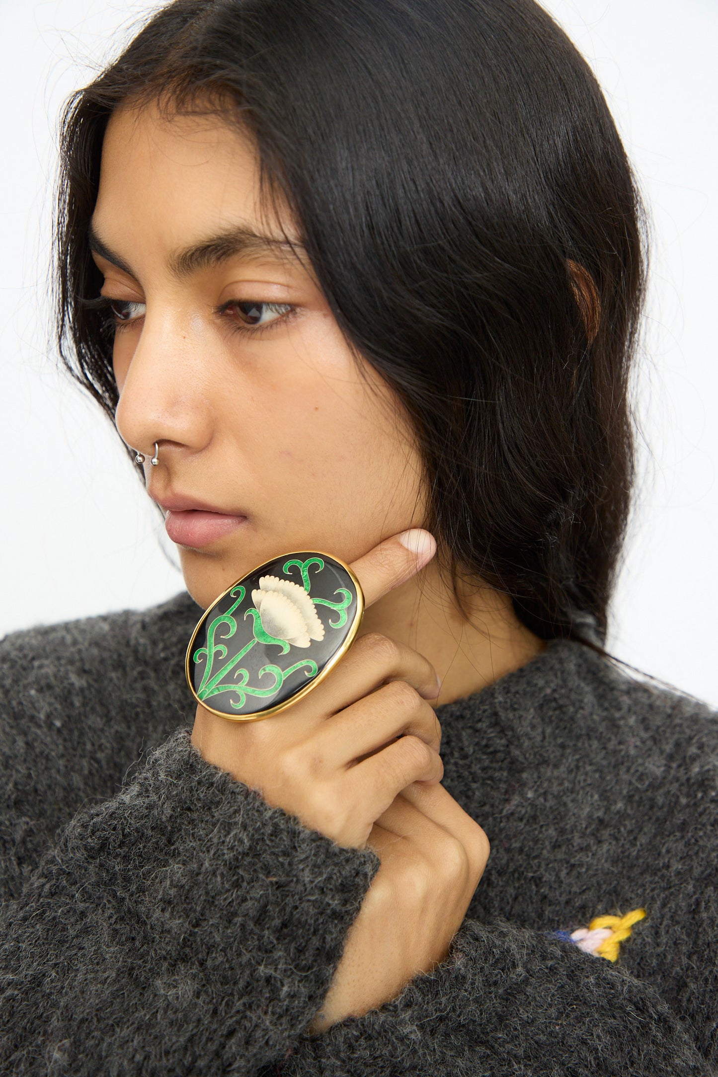 A person with long dark hair and a nose ring rests their chin on their hand, wearing the Sofio Gongli Oval Ring in Black with White Flower.