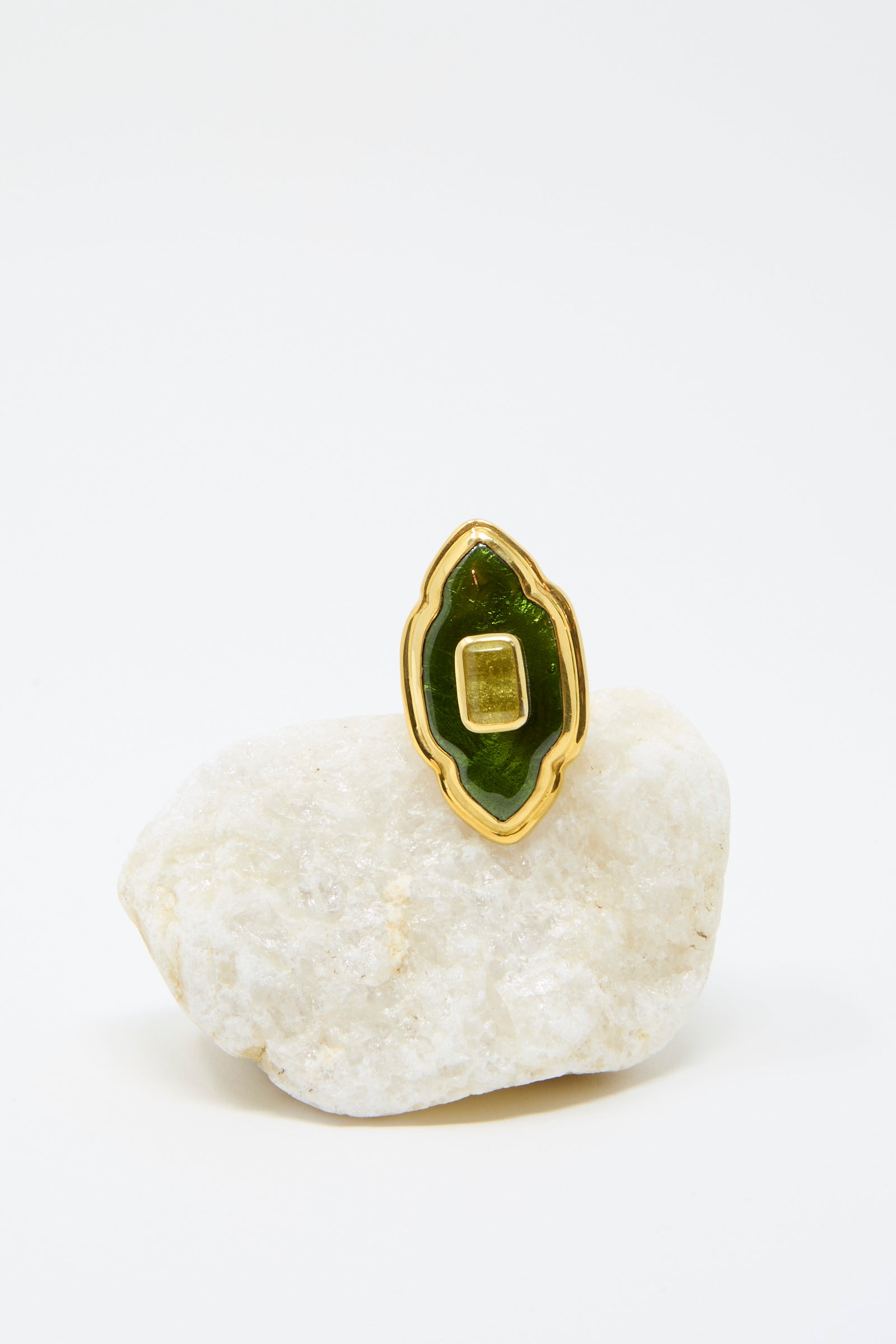 The Ring in Green and Gold by Sofio Gongli is a stunning piece, featuring a rectangular green stone with a gold border, sitting on a white stone against a plain backdrop. 