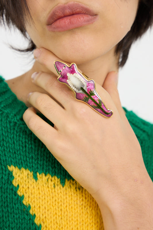 A person wearing a green and yellow sweater is photographed from the nose down, showcasing a Sofio Gongli ring in Purple Alligator with Flower on their finger.
