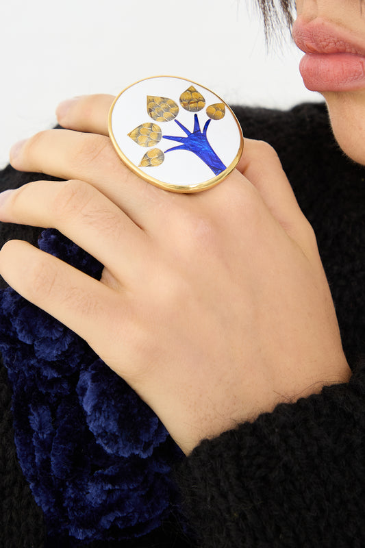 A close-up image of a person's hand wearing a Sofio Gongli Ring in Tree with a blue and gold Byzantine cloisonné design, against a black textured garment.