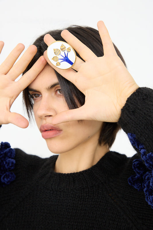 A person with short dark hair is posing with their hands framing one eye, showcasing a Sofio Gongli Ring in Tree with a blue and gold Byzantine cloisonné design worn on their finger. They are wearing a black sweater.