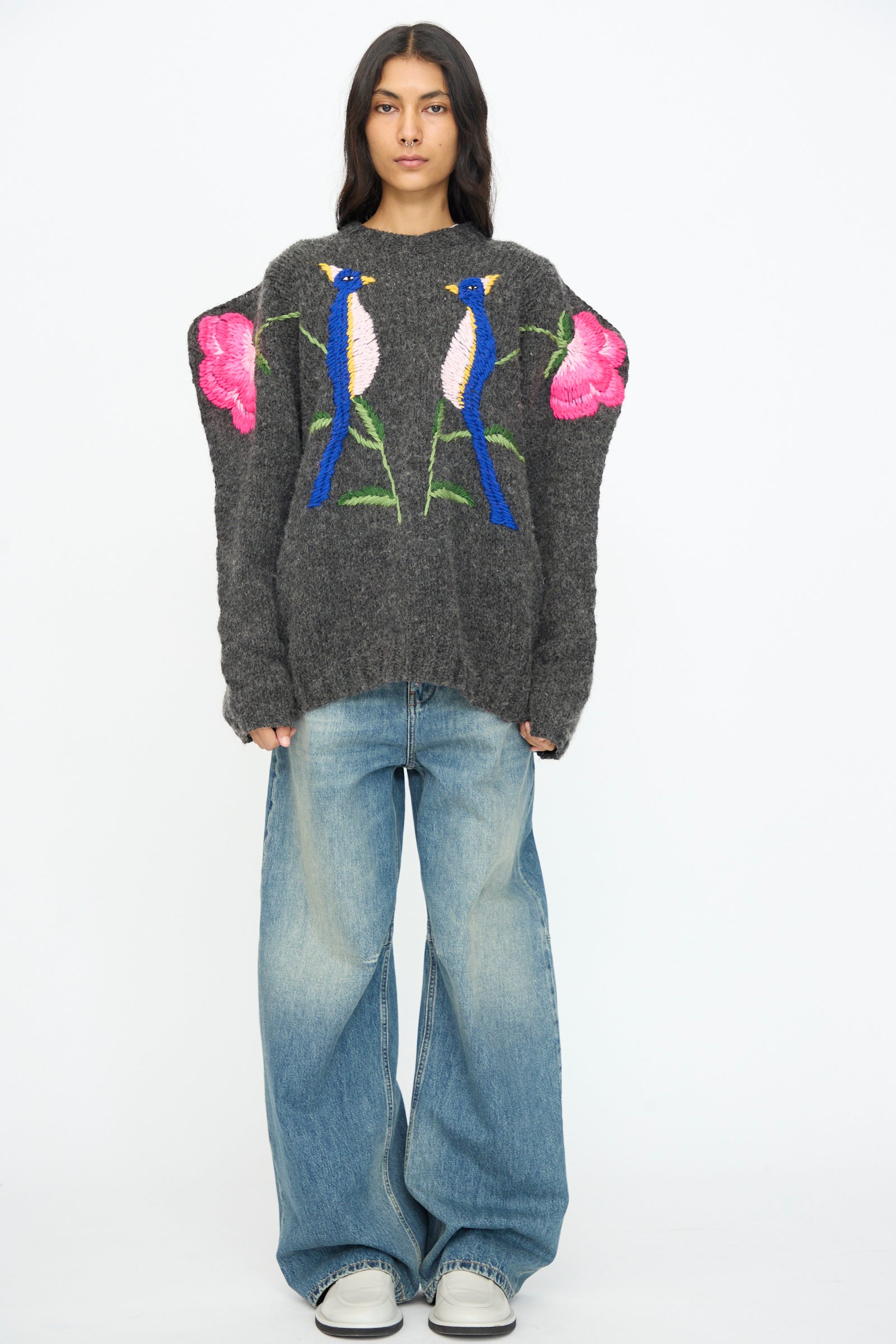 A person wears the Sofio Gongli Sweater in Charcoal with Birds and Flowers, paired with loose blue jeans and white sneakers, standing against a plain white background.