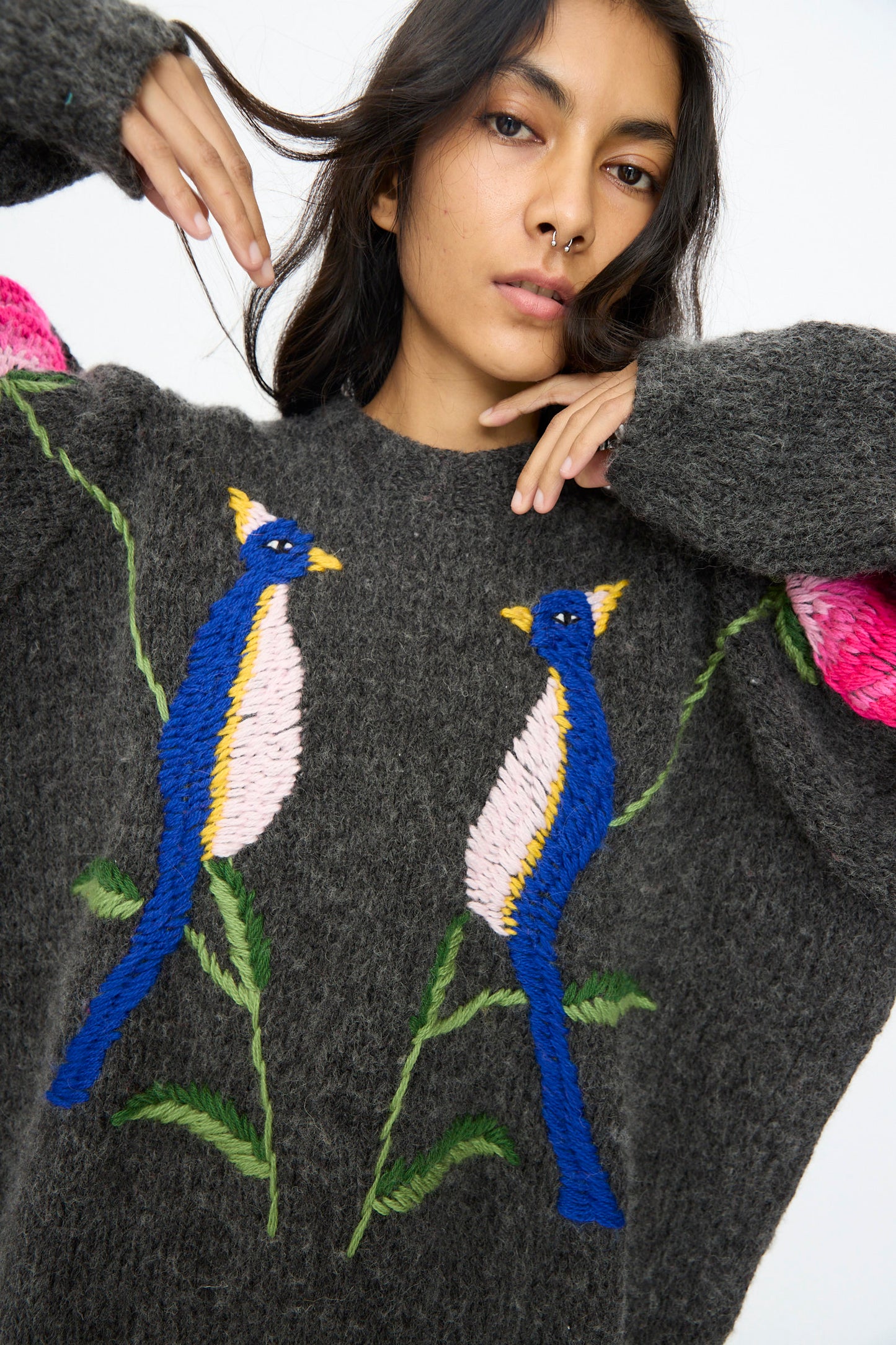 A woman wearing the Sofio Gongli Sweater in Charcoal with Birds and Flowers raises her arms near her face; the sweater features hand-embroidered colorful birds and green leaves on the front.