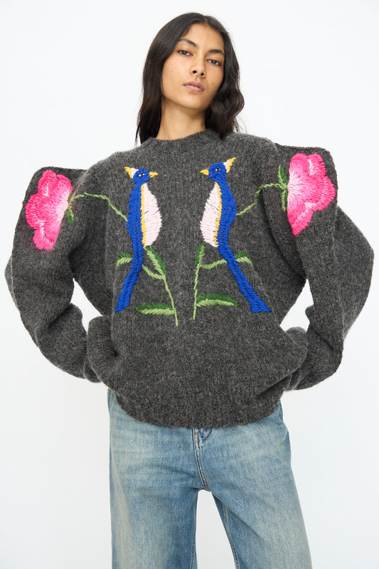 A person wears the Sofio Gongli Sweater in Charcoal with Birds and Flowers, featuring a hand-knit mohair wool blend with large puffed sleeves, hand-embroidered blue birds and pink flowers, paired with light blue jeans.