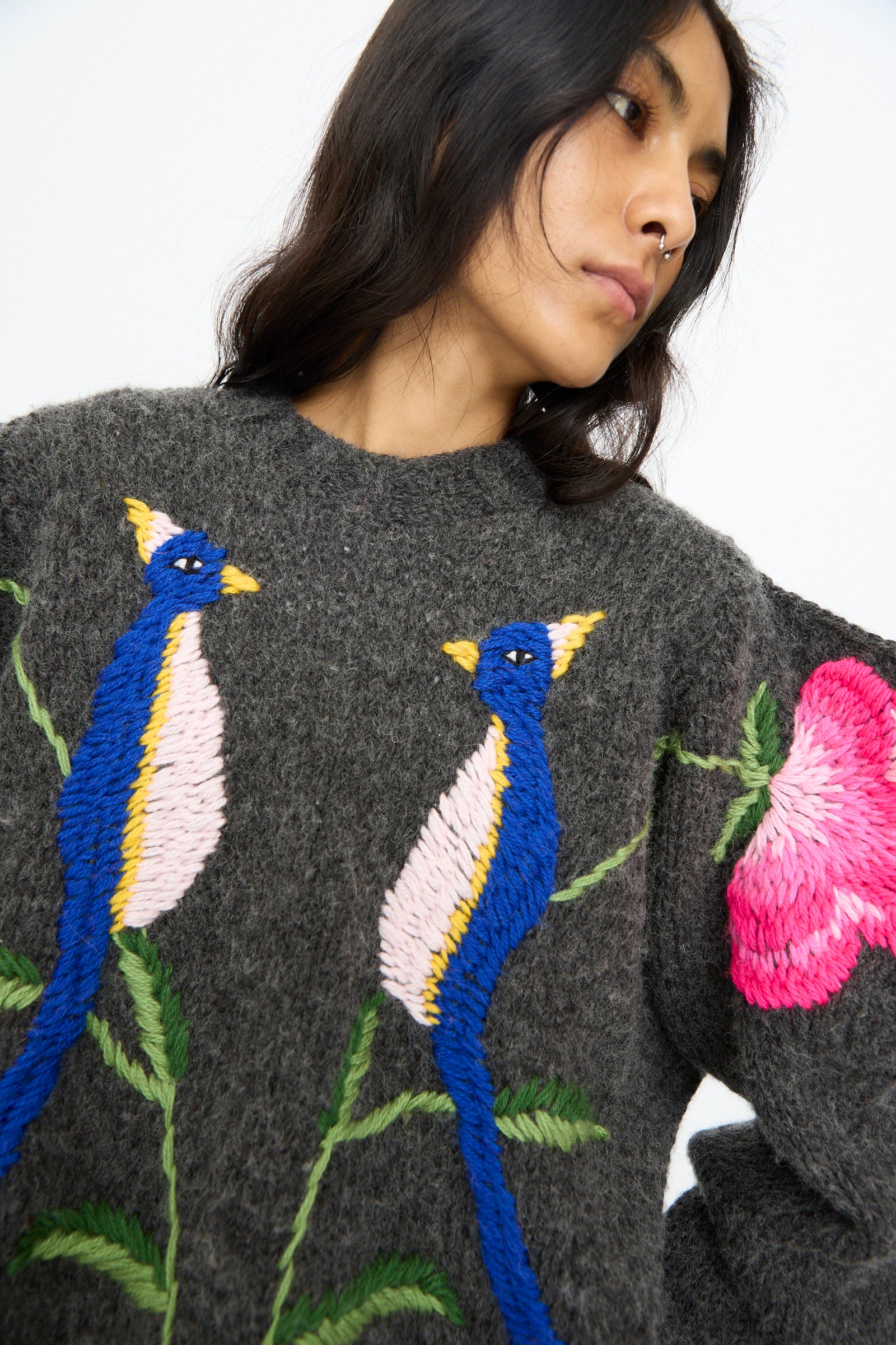 A person with long dark hair wears the Sofio Gongli Sweater in Charcoal with Birds and Flowers, a hand-knit gray sweater featuring large embroidered blue birds and a pink flower.
